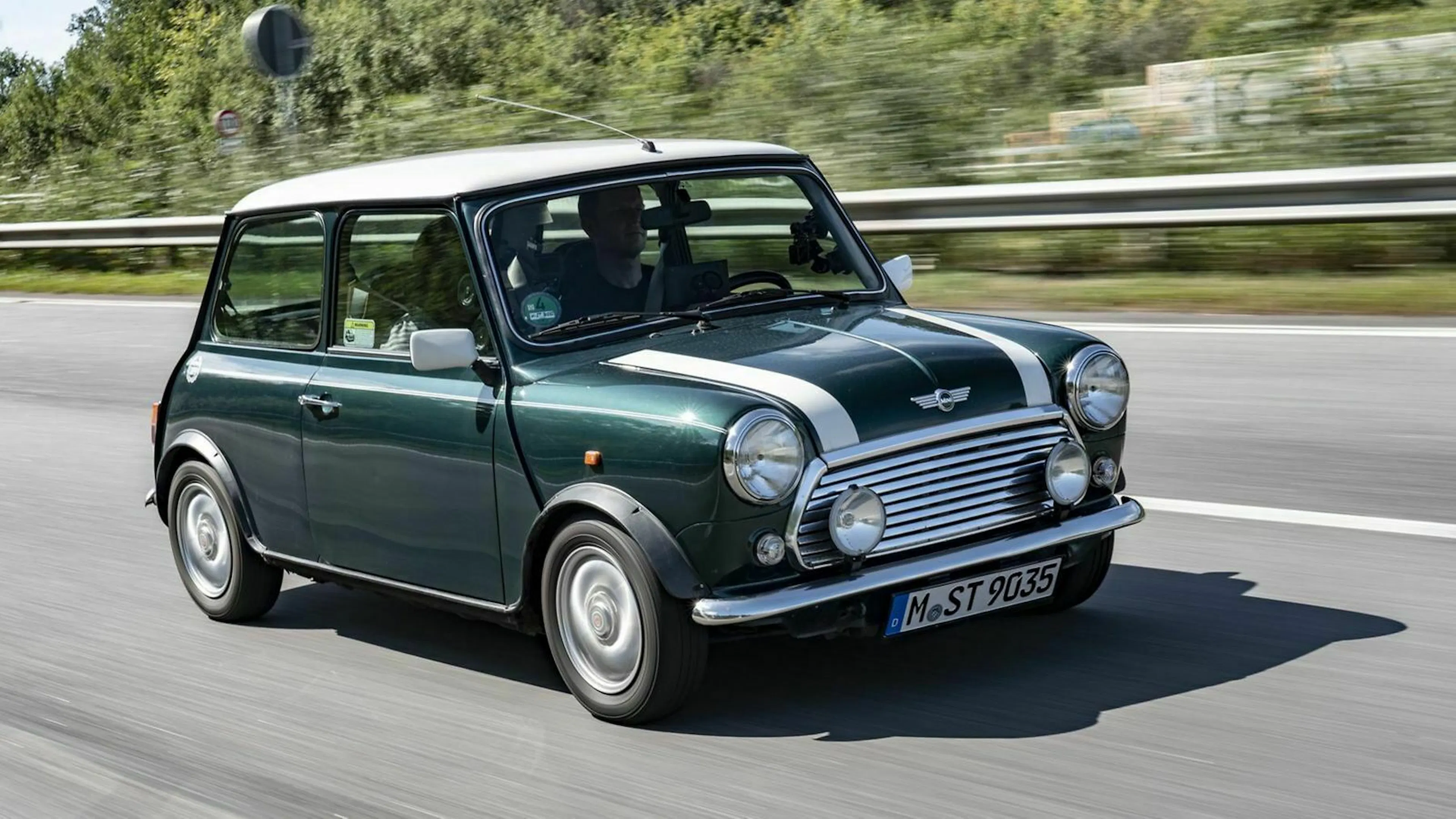 Grüner klassischer Mini Cooper mit weißem Dach und Rennstreifen fährt auf einer Autobahn, von Bäumen im Hintergrund gesäumt.