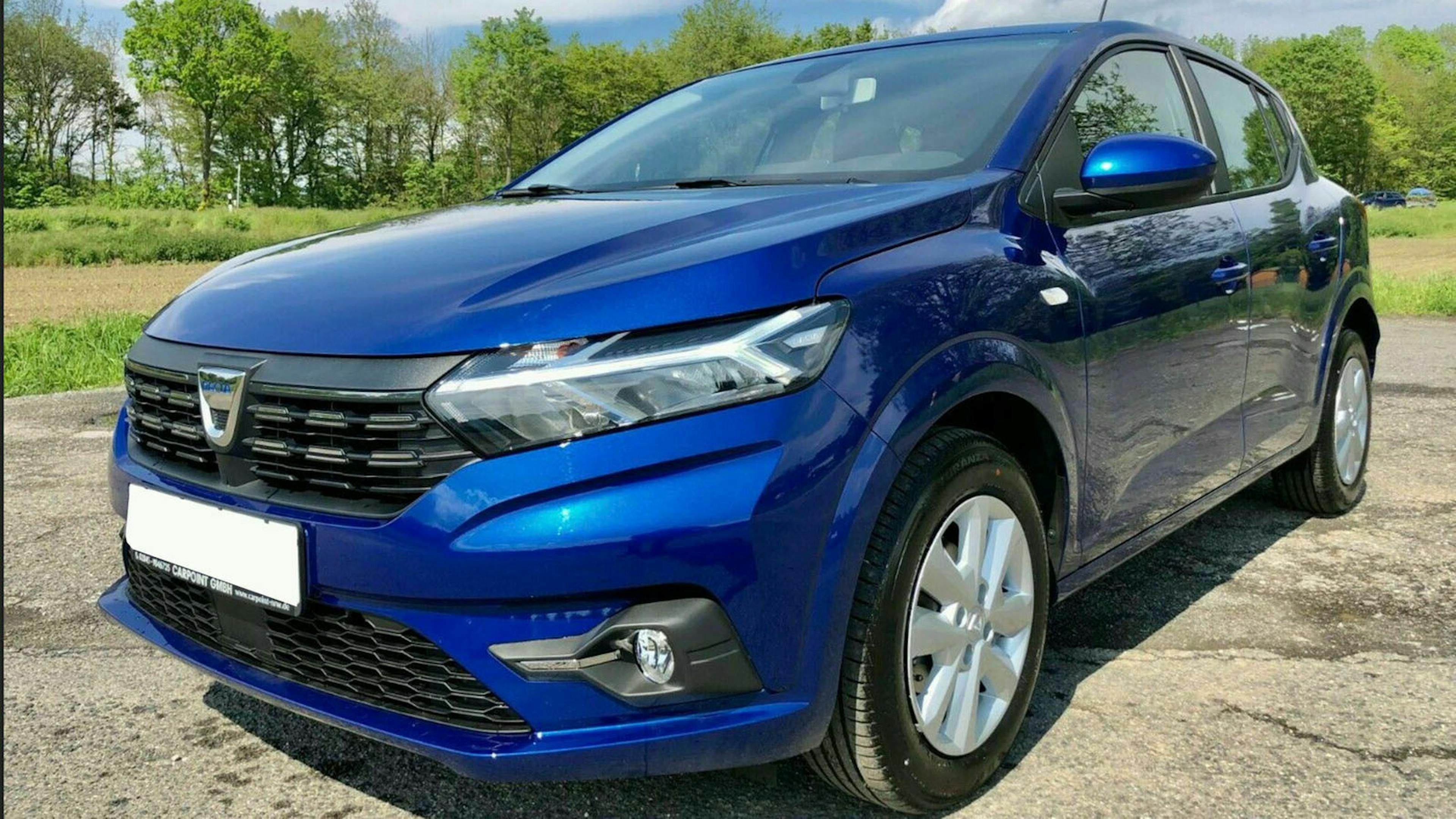 Ein blauer Dacia Sandero steht auf einem Parkplatz am Waldrand.