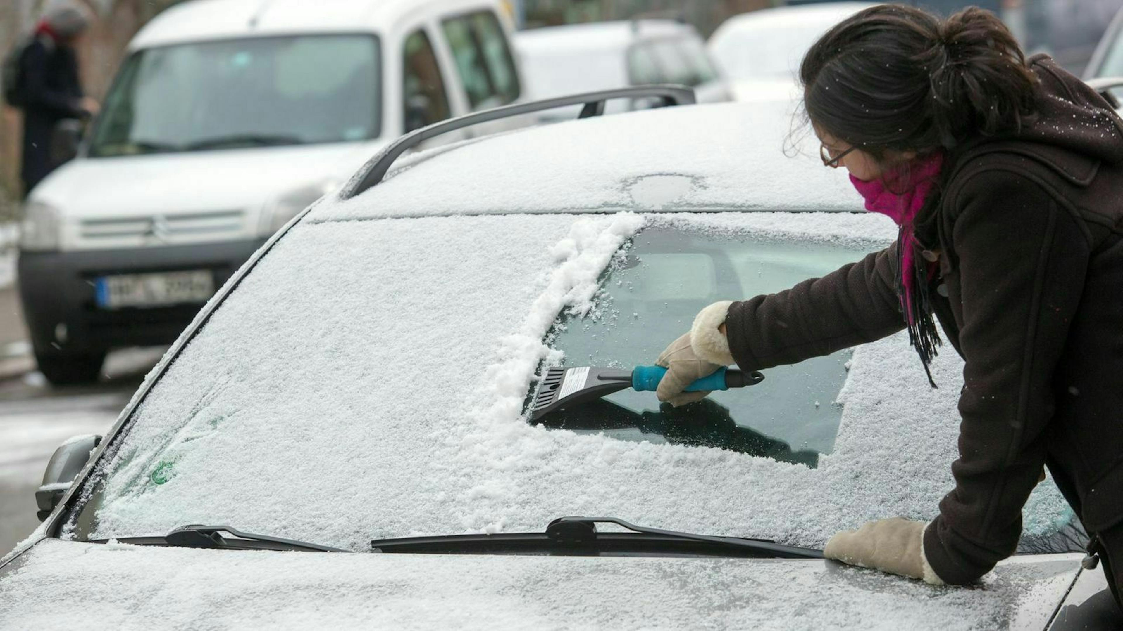 Zu sehen ist eine Frau die die Frontscheibe eines Autos enteist 
