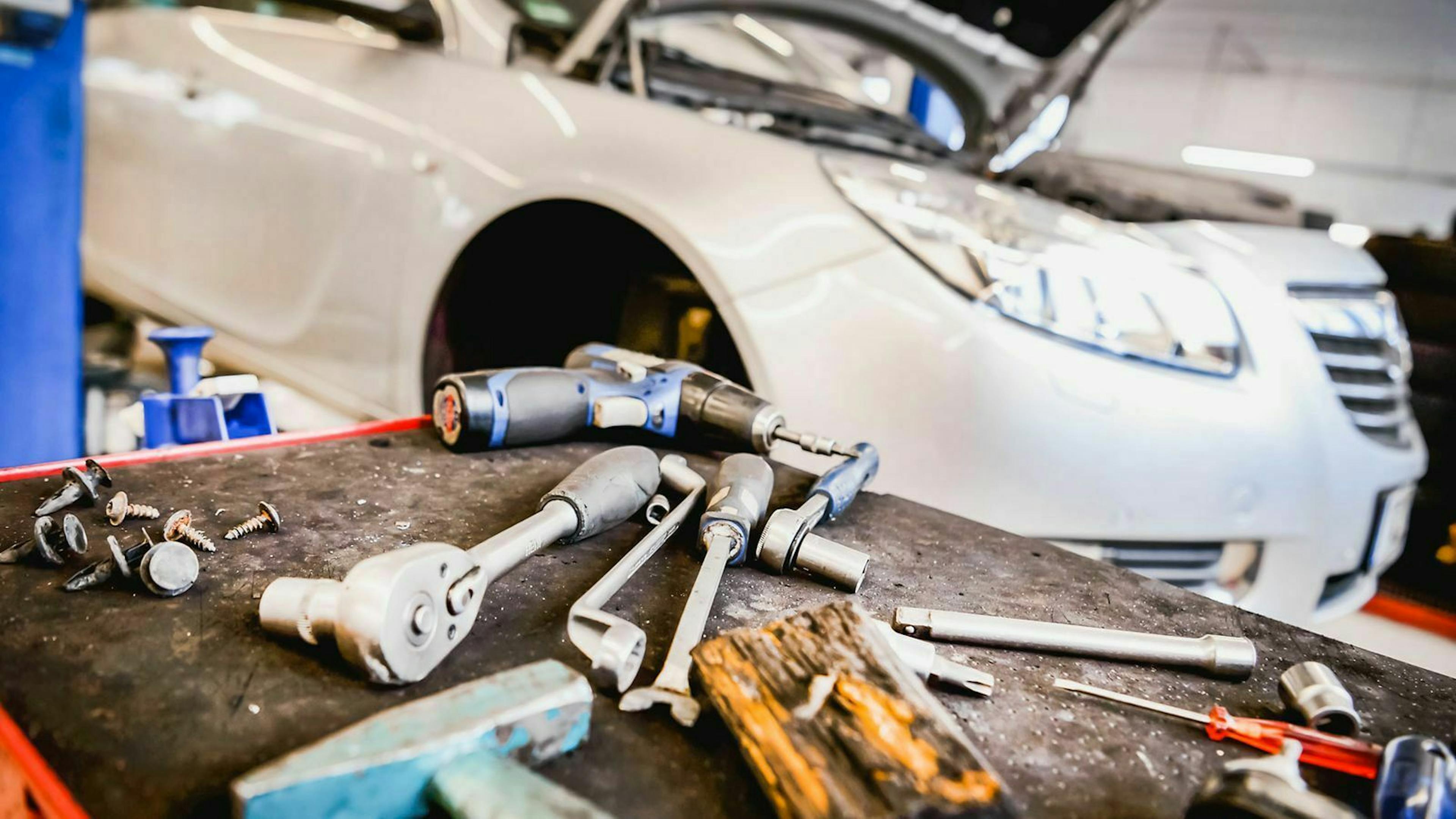 Im Vordergrund liegen Werkzeuge während unscharf im Hintergrund ein Auto mit geöffneter Motorhaube steht.