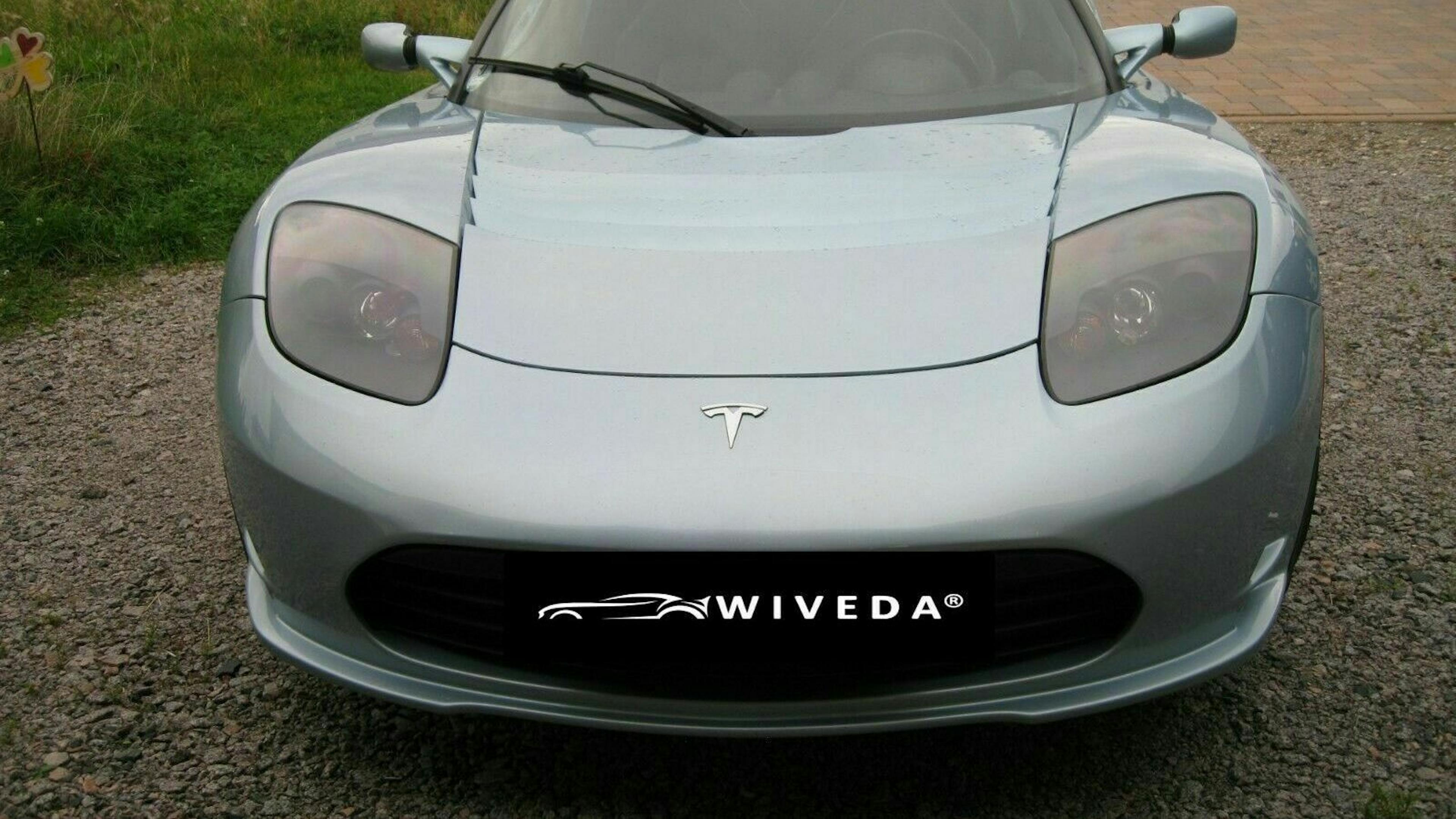 Ein silberner Tesla Roadster steht auf einem Parkplatz vor einer Wiese