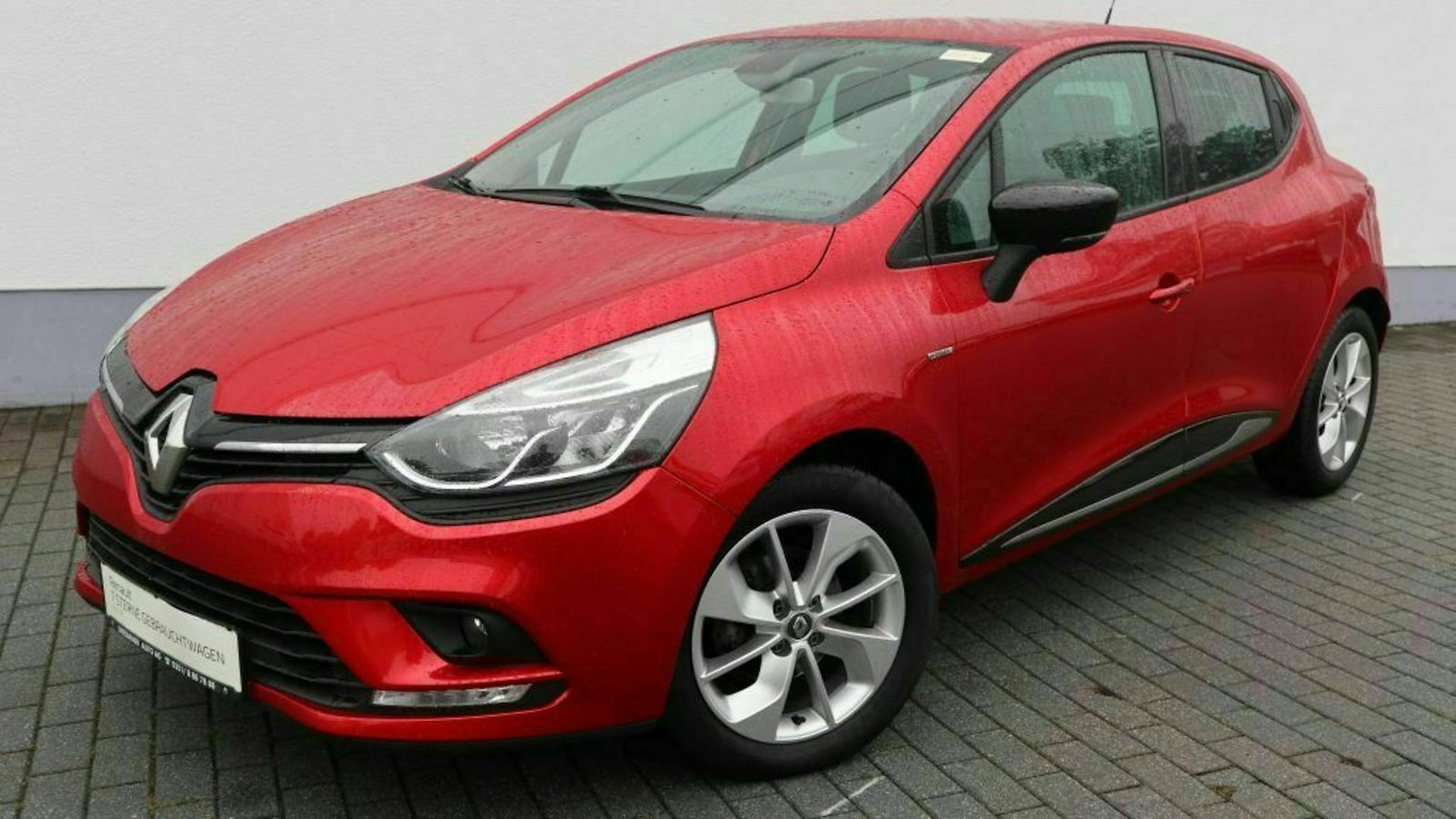 Rotes Auto der Marke Renault, geparkt auf gepflastertem Boden vor einer weißen Wand, Vorderansicht mit Fokus auf Scheinwerfer und Frontstoßstange.