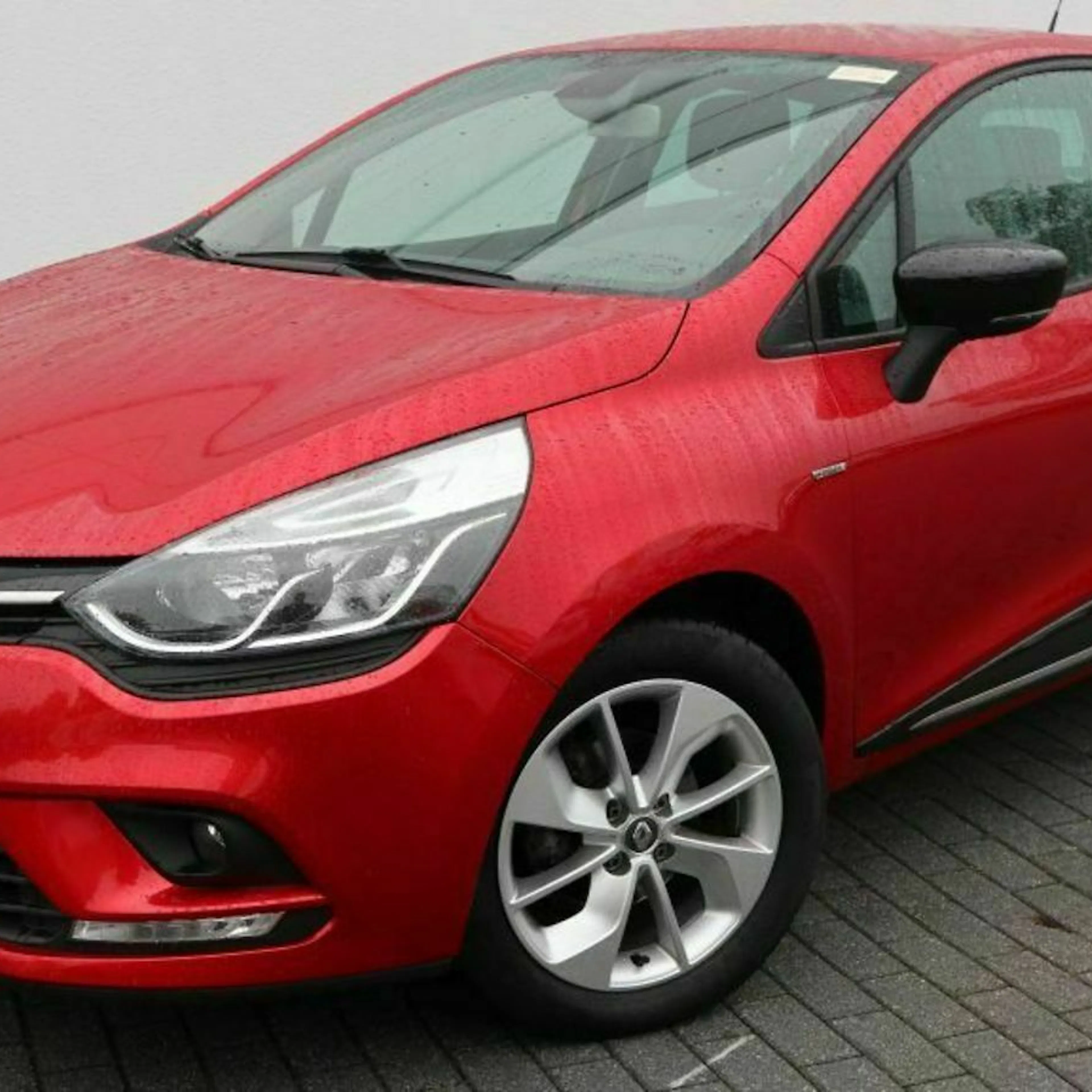 Rotes Auto der Marke Renault, geparkt auf gepflastertem Boden vor einer weißen Wand, Vorderansicht mit Fokus auf Scheinwerfer und Frontstoßstange.