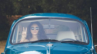 Eine junge Frau mit Sonnenbrille sitzt in einem blauen Volkswagen, mit der rechten Hand zeigt sie ein Peace-Zeichen