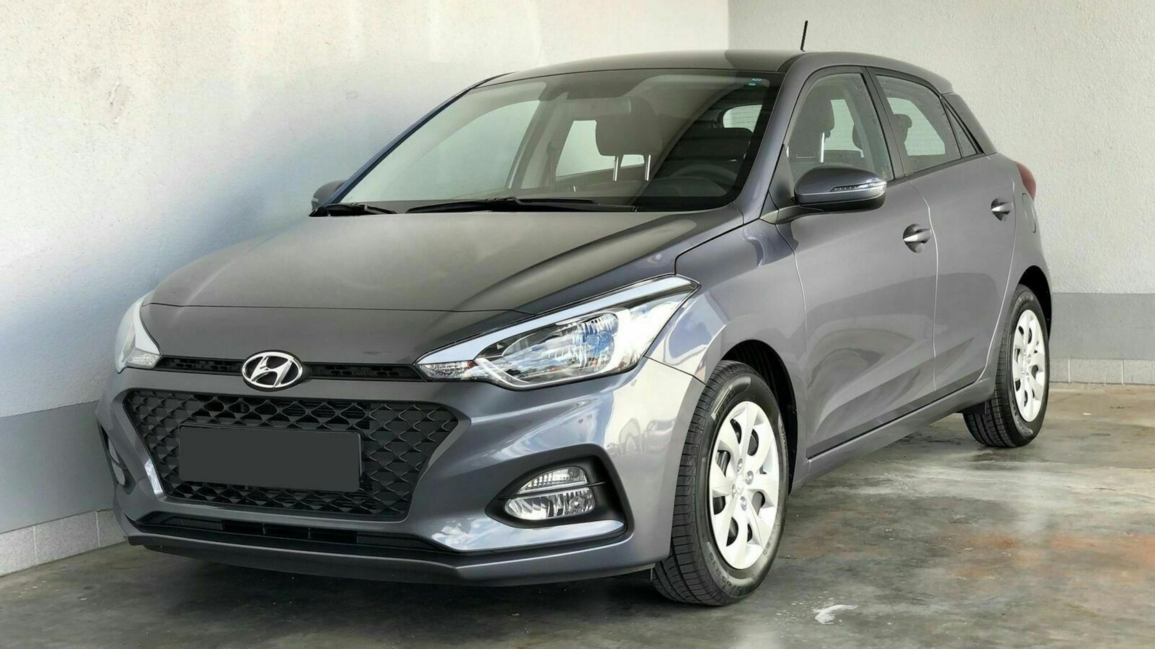 Grauer Hyundai-Kleinwagen parkt in einer Garage. Vorderansicht zeigt markanten Kühlergrill und weiße Felgen auf Betonboden.