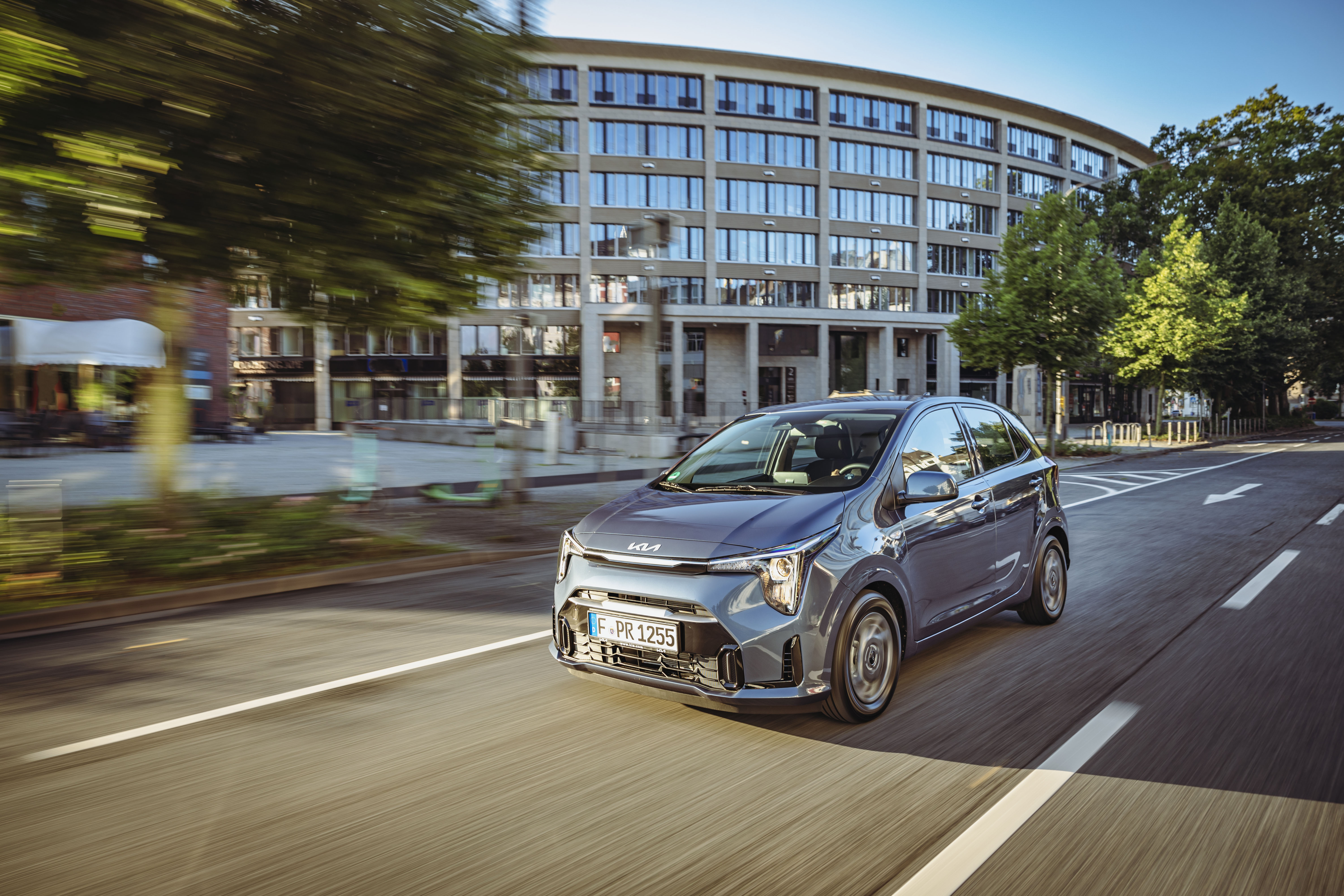 Blaugrauer Kia Picanto fährt auf einer Stadtstraße an einem großen Bürogebäude mit runder Fassade vorbei.