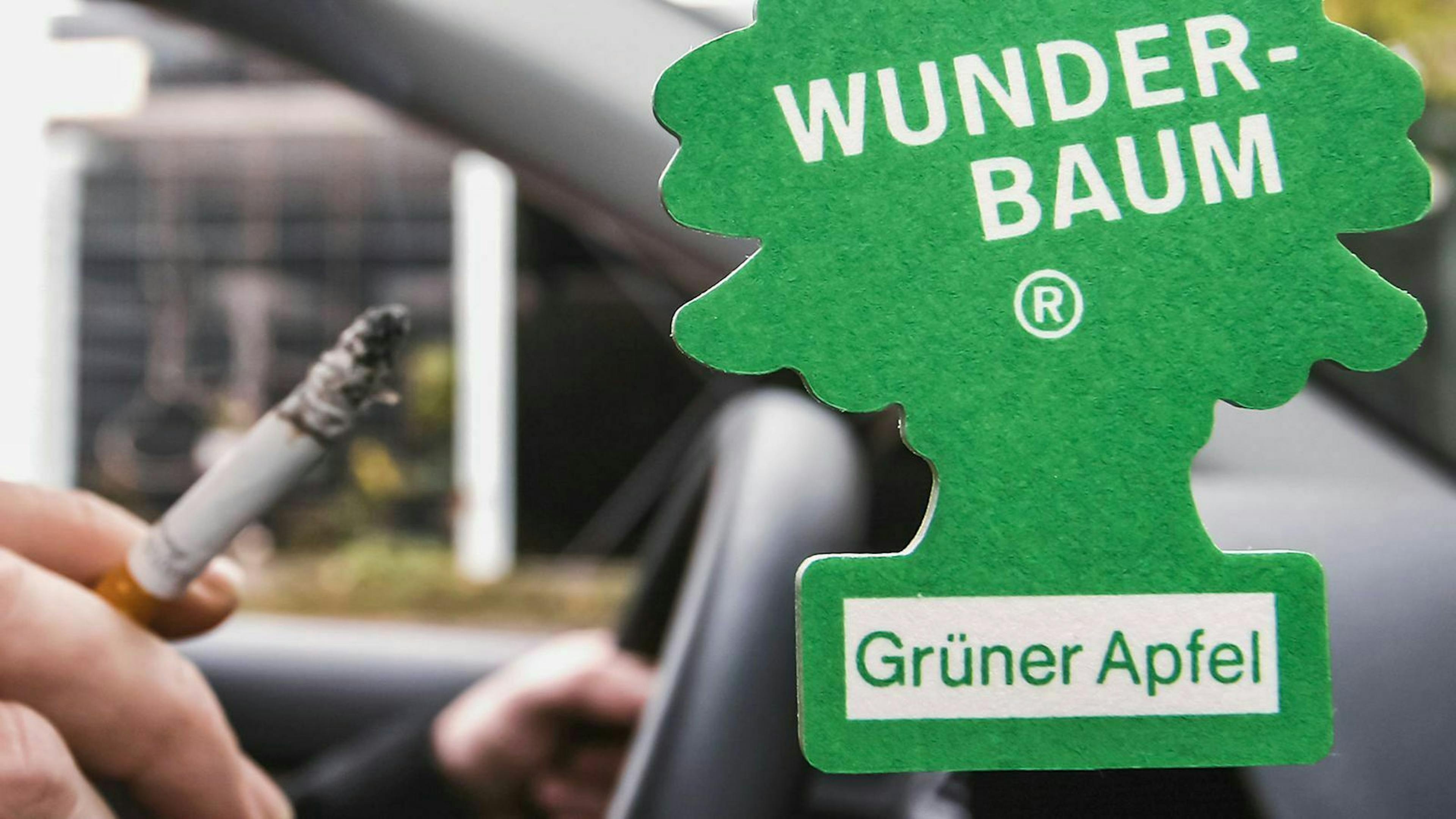 Ein "Wunderbaum" hängt am Rückspiegel eines Autos während der Fahrer im Hintergrund eine Zigarette hält.
