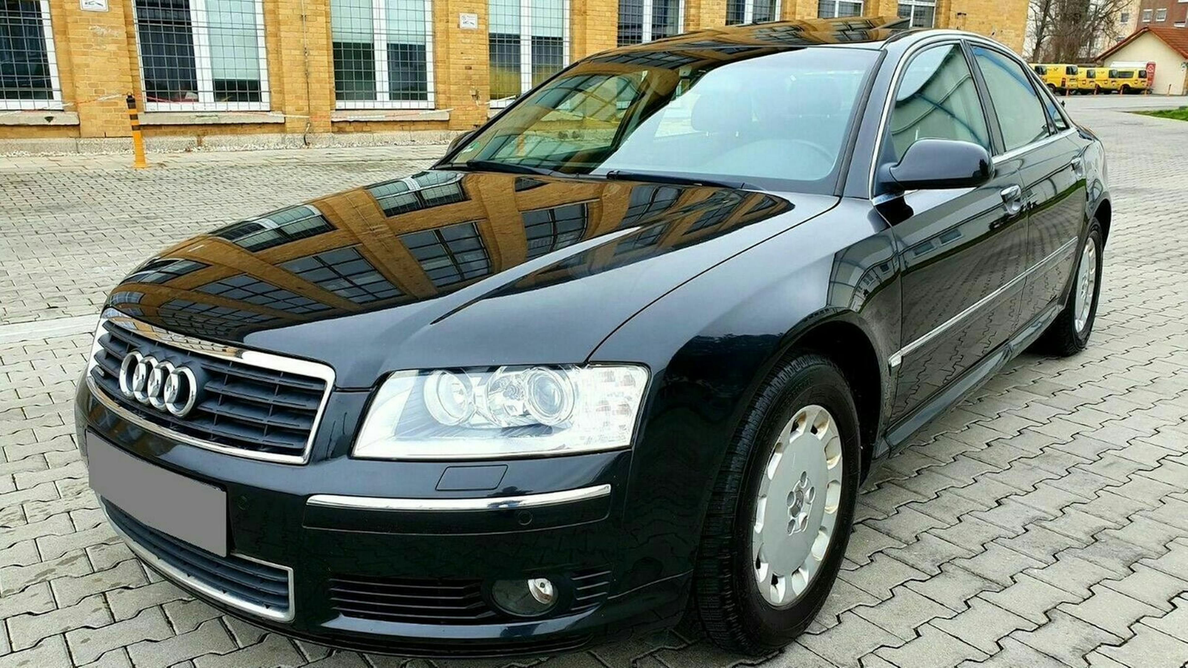 Schwarzer Audi-Limousine auf einem gepflasterten Parkplatz vor einem Backsteingebäude. Vorderansicht mit spiegelnder Karosserie.
