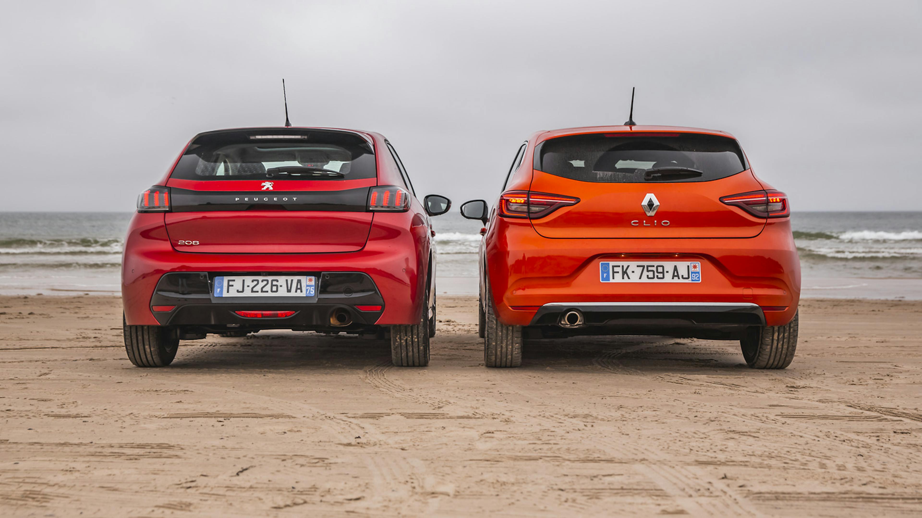 Peugeot 208 und Renault Clio in der Heck-Ansicht. Stehend nebeneinander am Strand. Das Meer im Hintergrund