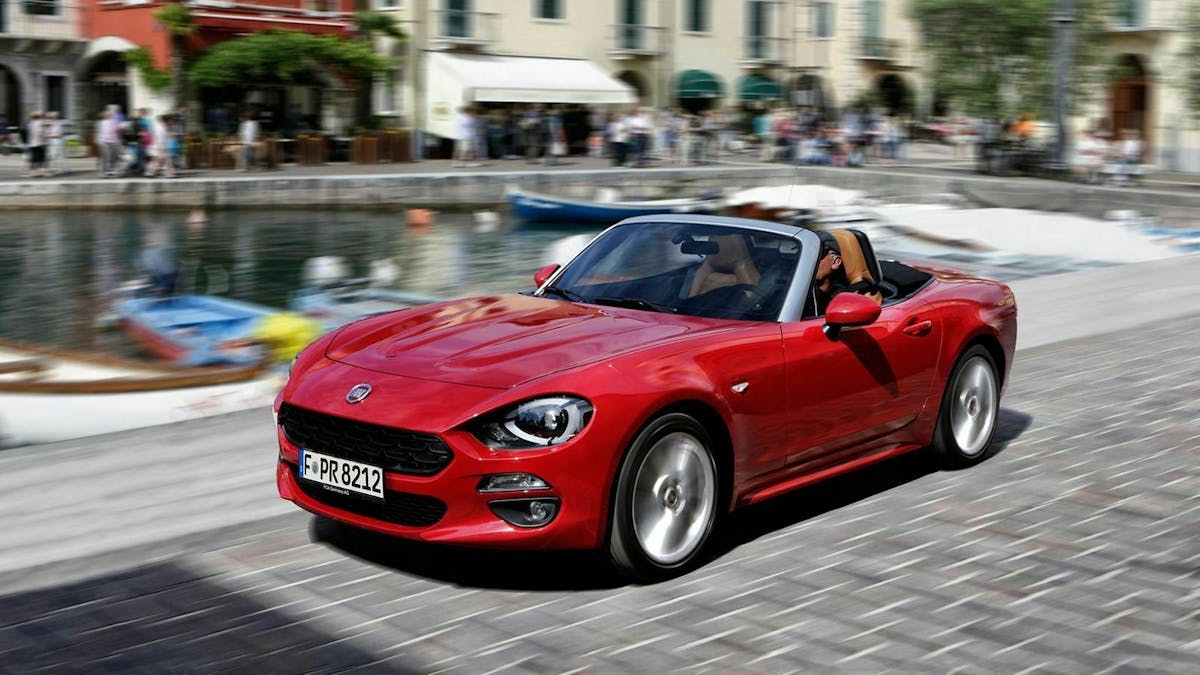 Roter Fiat 124 Spider fahrend, in der Ansicht von vorne-links. Geschäftiges Treiben an einem Hafenbecken im Hintergrund