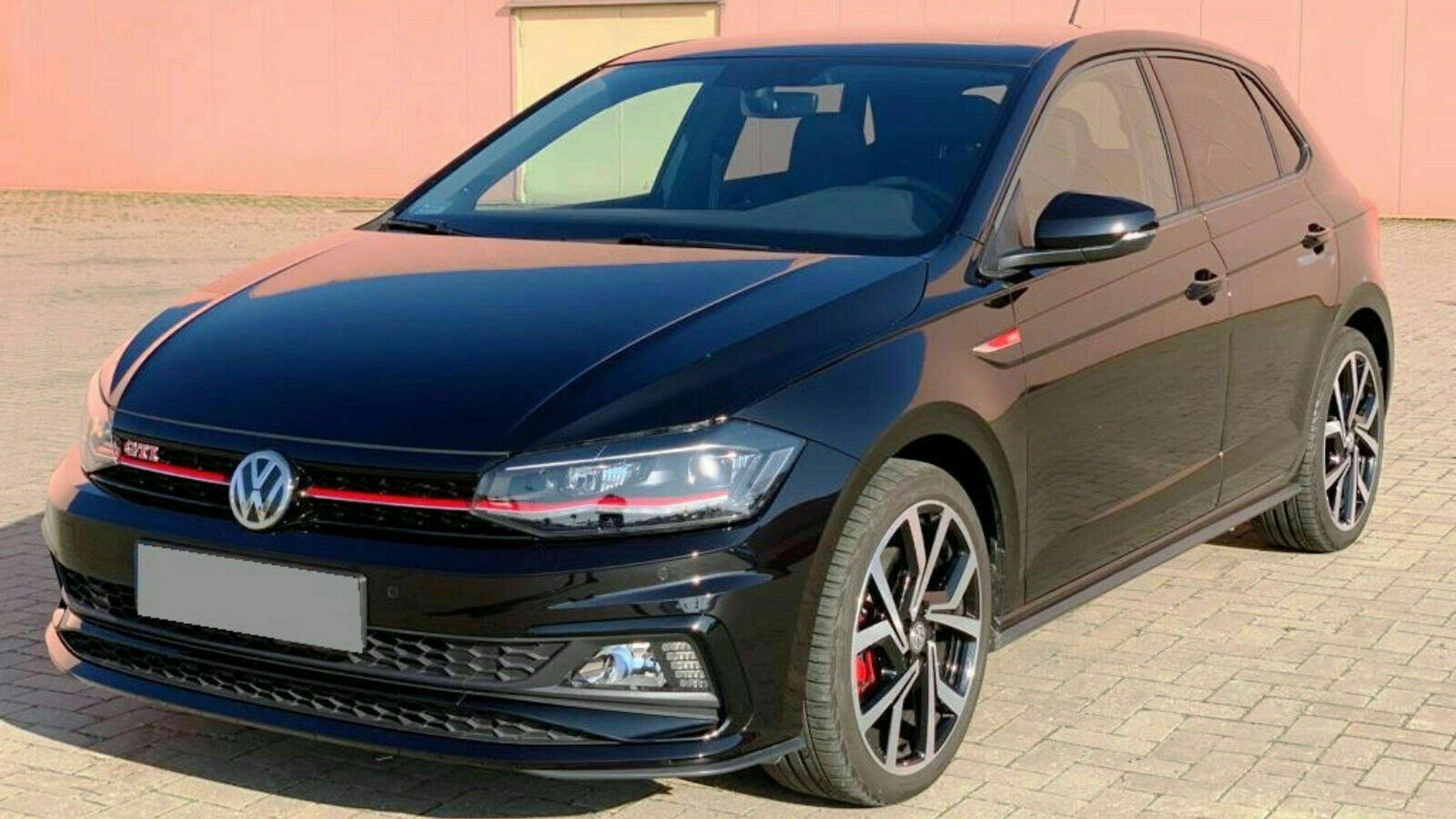 Schwarzer VW Polo GTI, geparkt auf einem Pflastersteinboden vor einer rosafarbenen Wand. Vorderansicht, erkennbar an roten Akzenten.