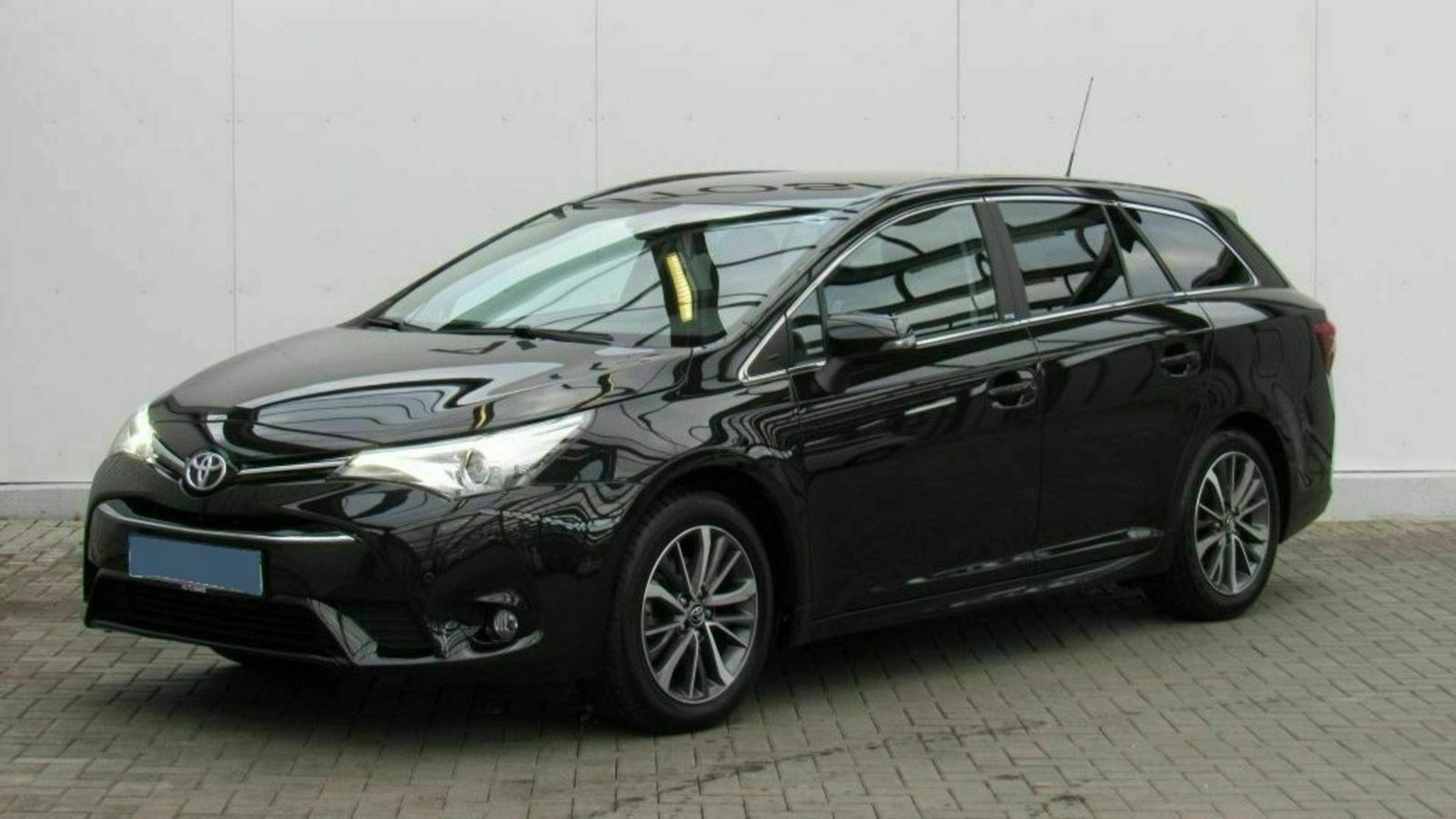 Schwarzer Kombi steht auf grauem Pflaster vor neutraler Wand, seitlich von vorne fotografiert.