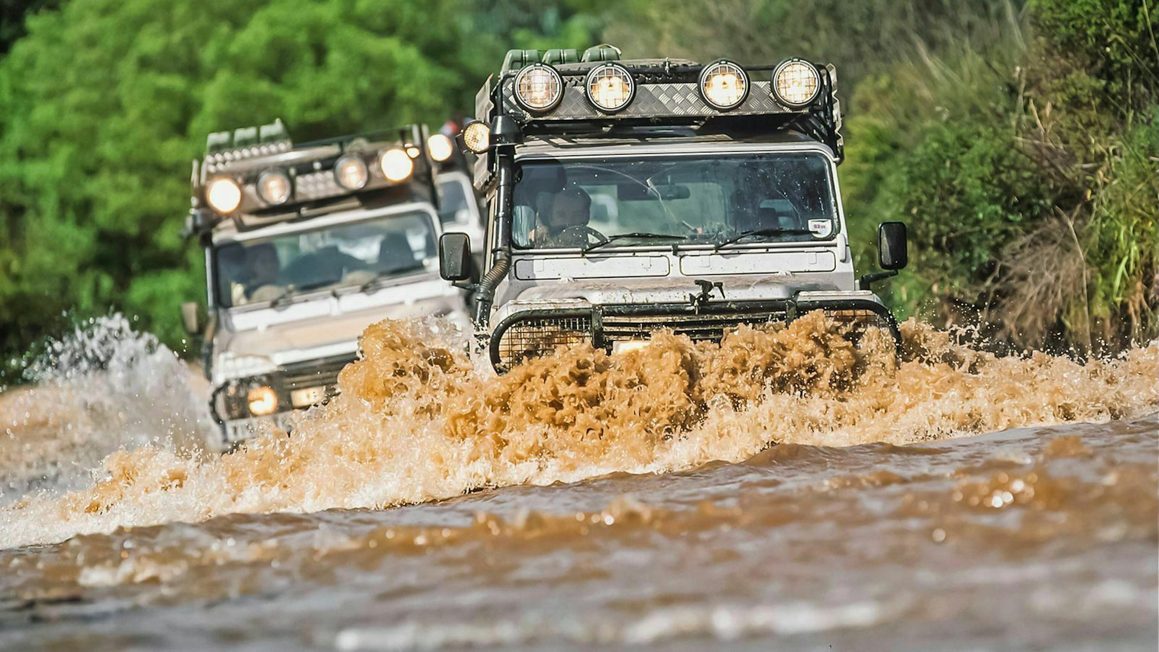 Zwei Land Rover Defender fahren durch einen Fluss während das Wasser bis zu den Scheinwerfern schwappt.