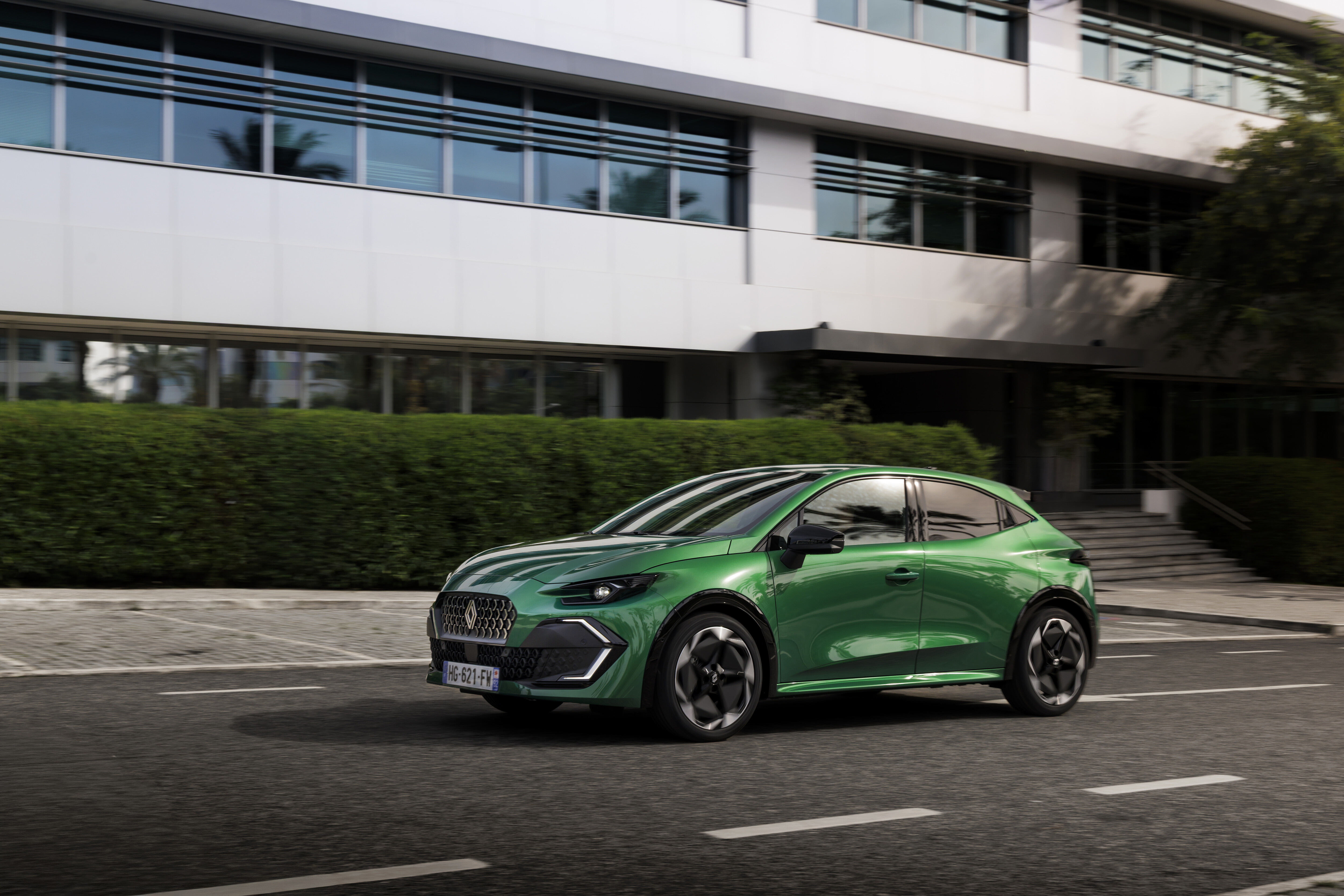Grüner Renault Clio Full Hybrid E-Tech in Fahrt vor einem Bürogebäude mit Hecke.