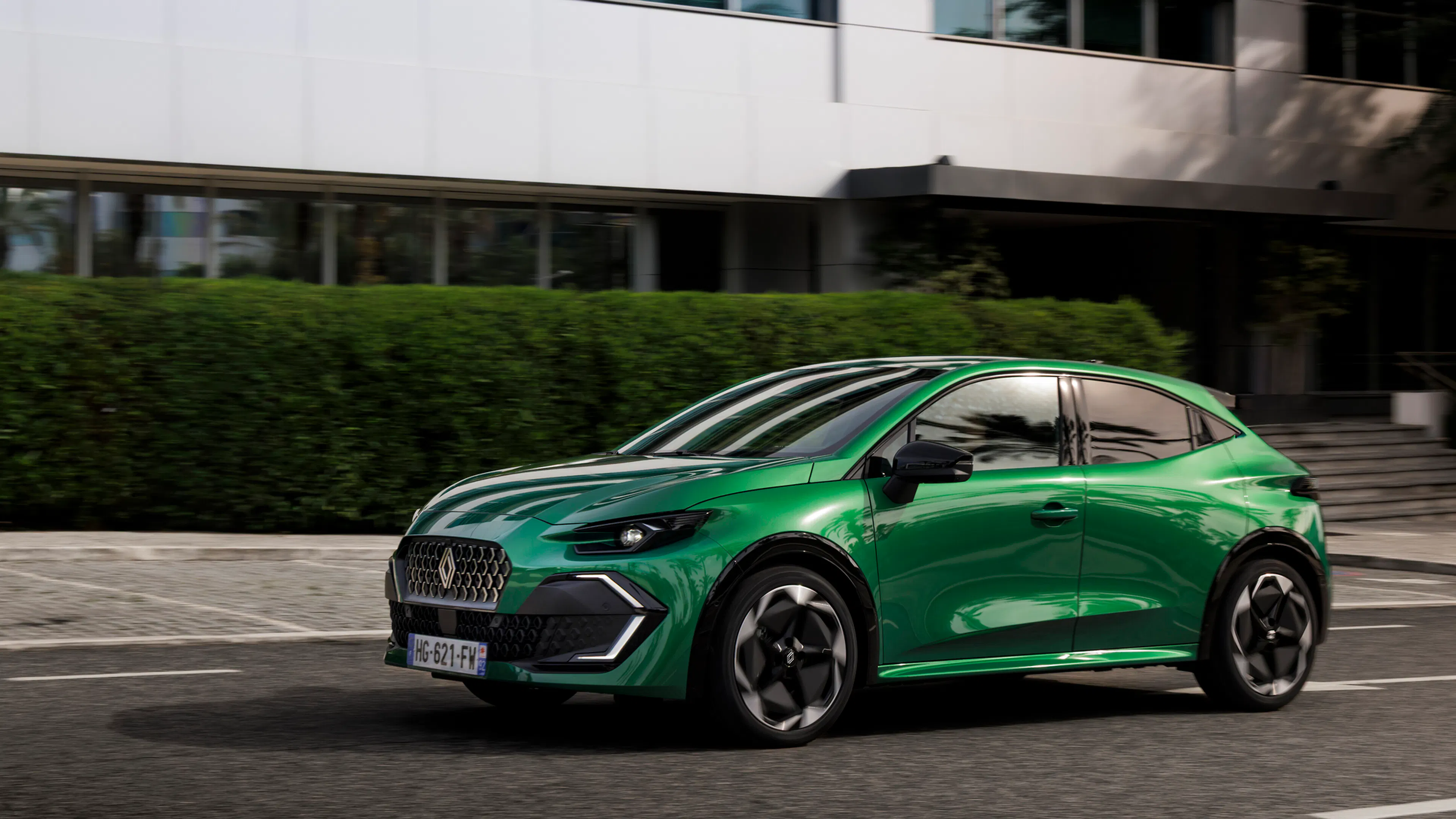 Grüner Renault Clio Full Hybrid E-Tech in Fahrt vor einem Bürogebäude mit Hecke.