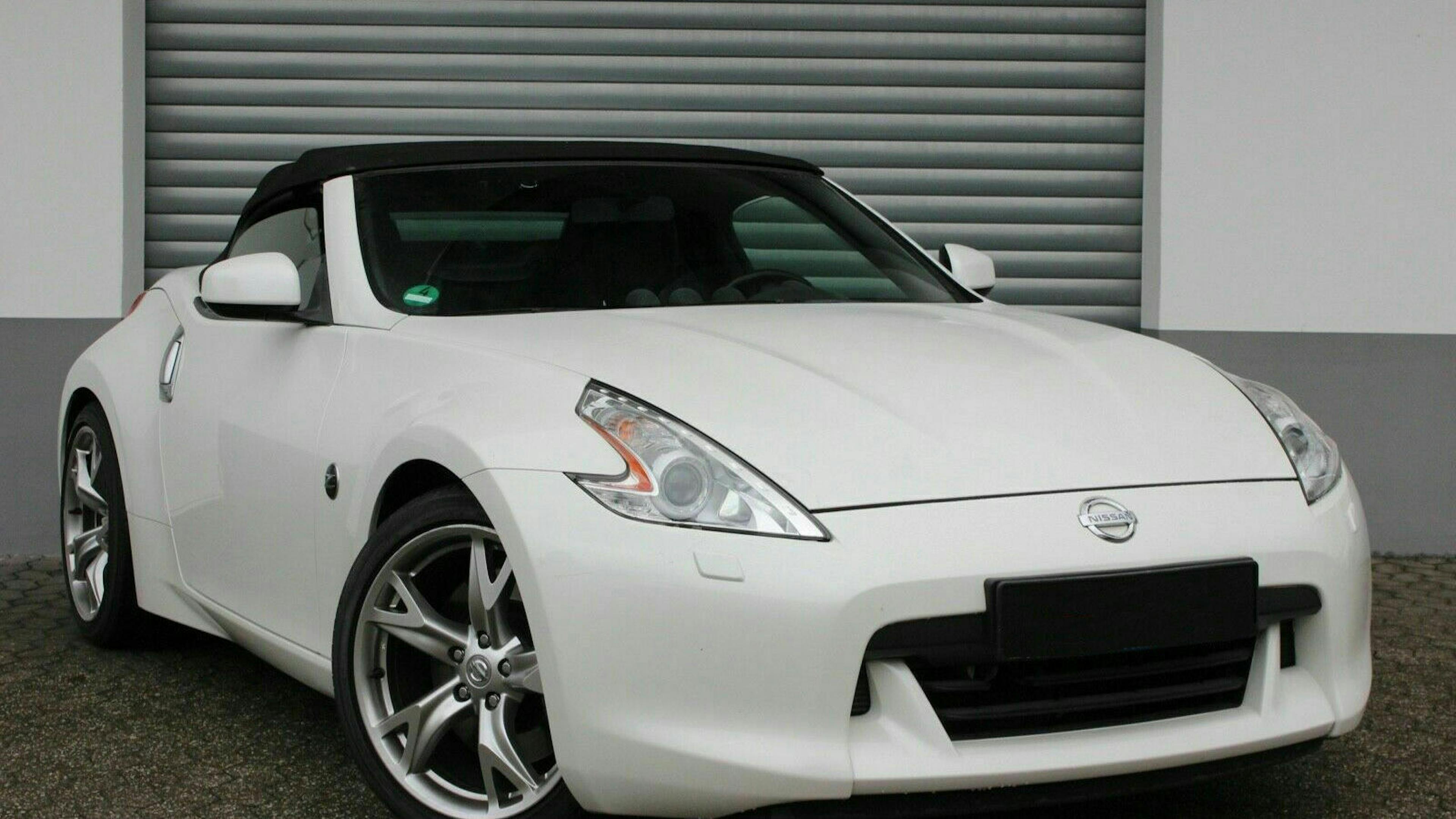 Ein weißer Nissan 370Z mit Heckantrieb steht vor einer weißen Wand mit grauem Rolltor
