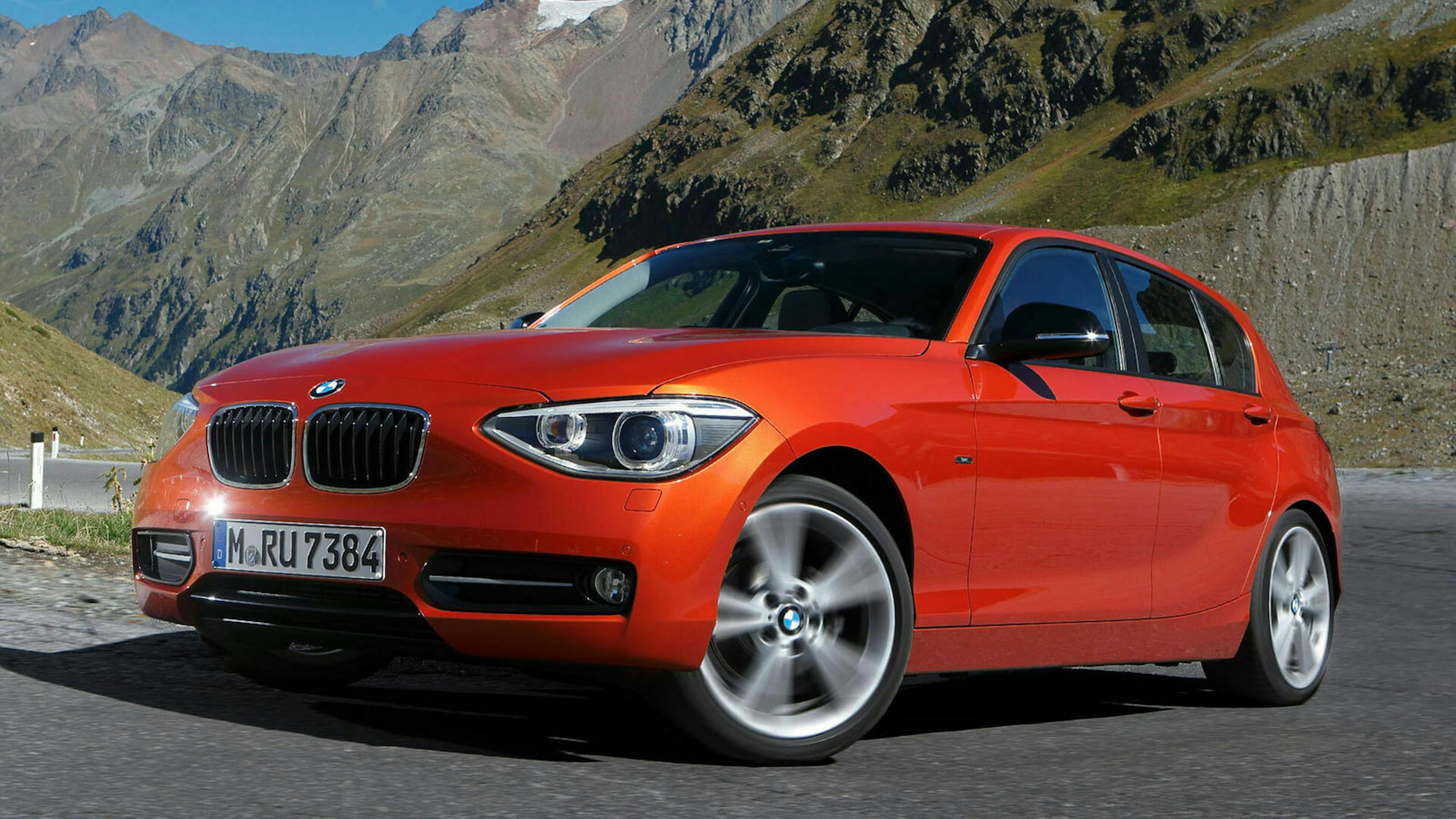 Ein roter BMW 1er mit Heckantrieb steht auf einer Straße vor einem Bergpanorama