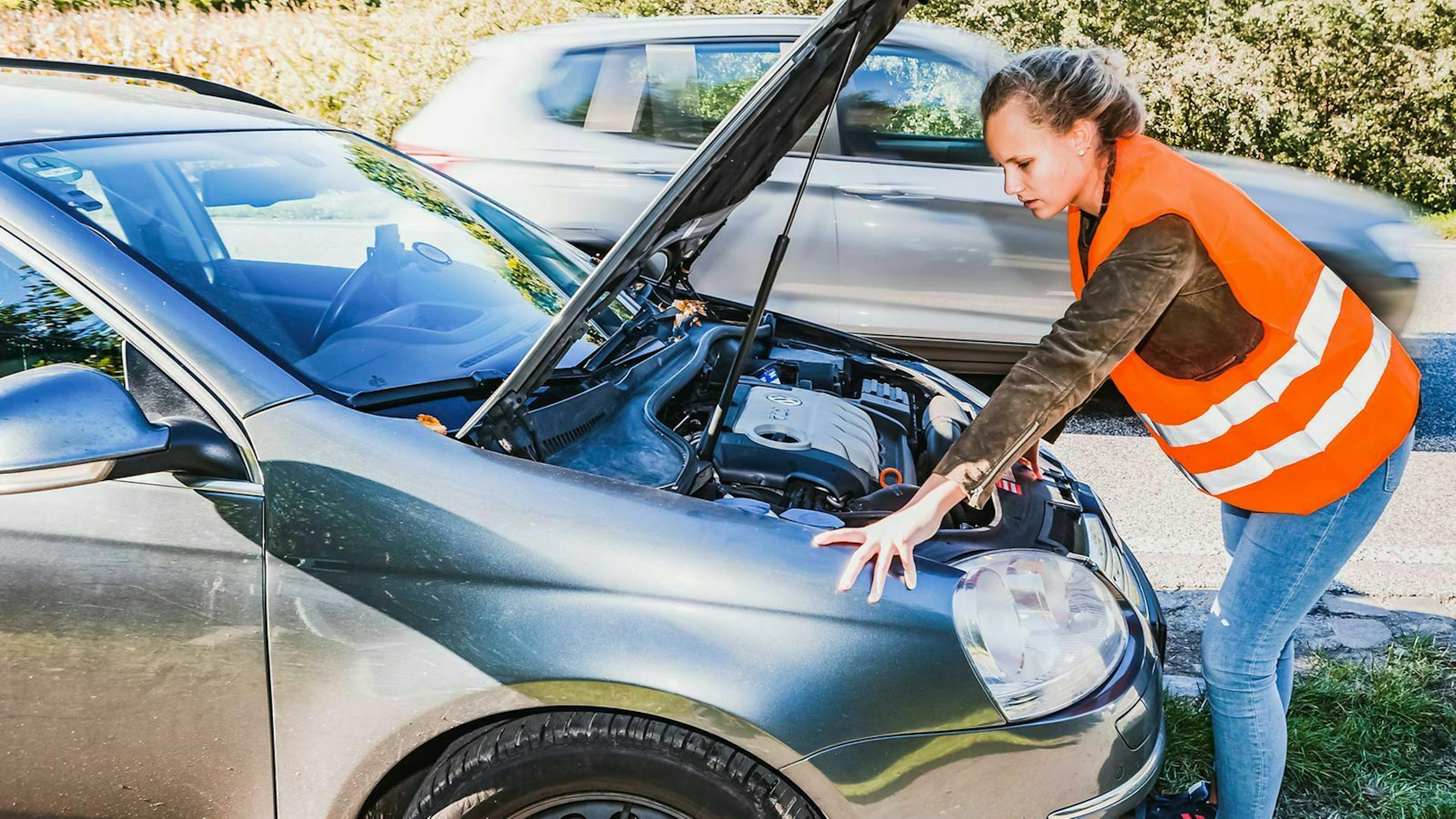 Eine Frau steht gekleidet mit einer Warnweste vor dem geöffneten Motorraum ihres Autos.