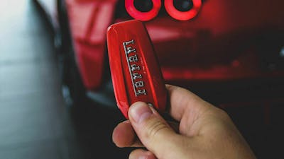 Eine Hand hält einen Autoschlüssel mit rotem Anhänger der Marke Ferrari vor dem unscharf erkennbaren Heck eines roten Ferraris