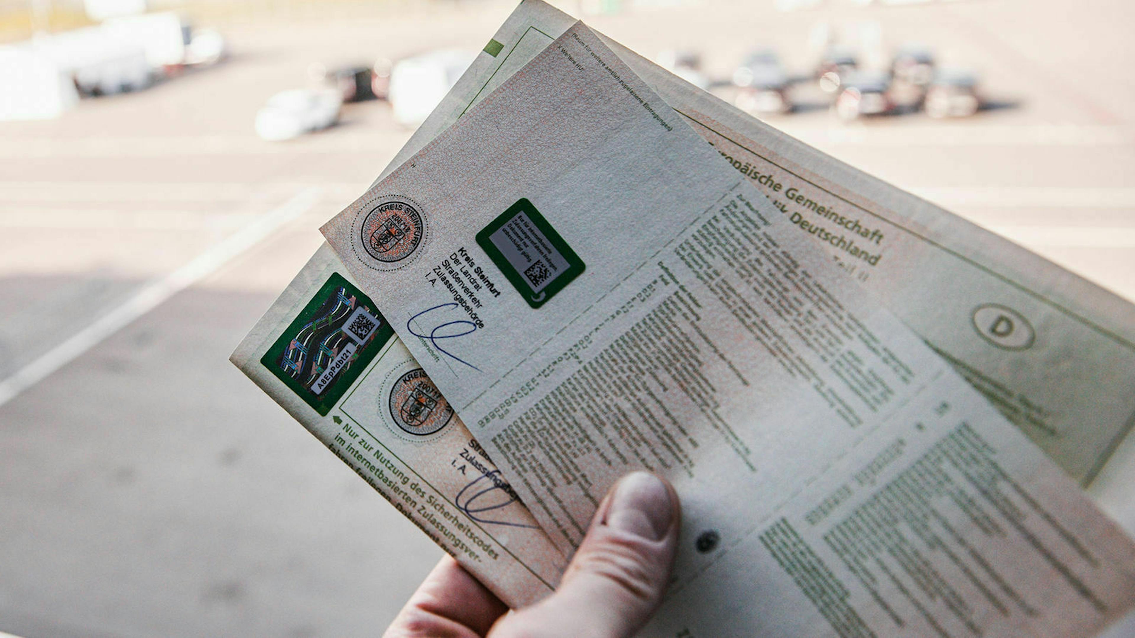 Jemand hält Fahrzeugpapiere in der Hand. Im Hintergrund ist ein Parkplatz zu sehen
