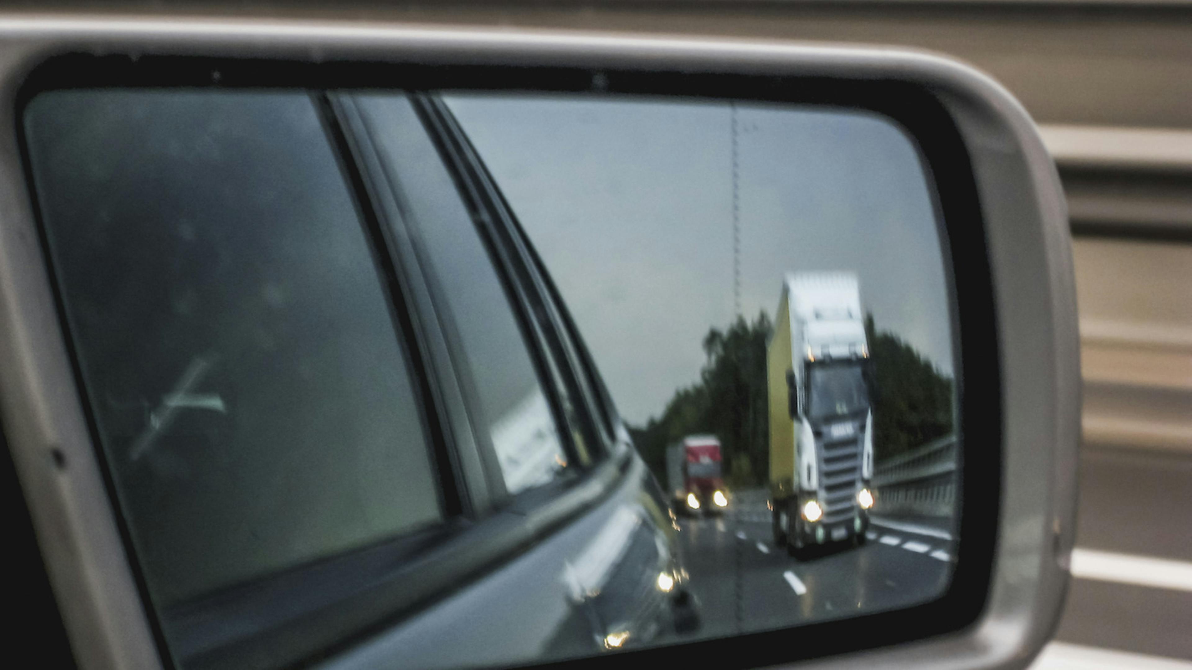 In einem PKW Rückspiegel ist in der Ferne ein LKW zu sehen.