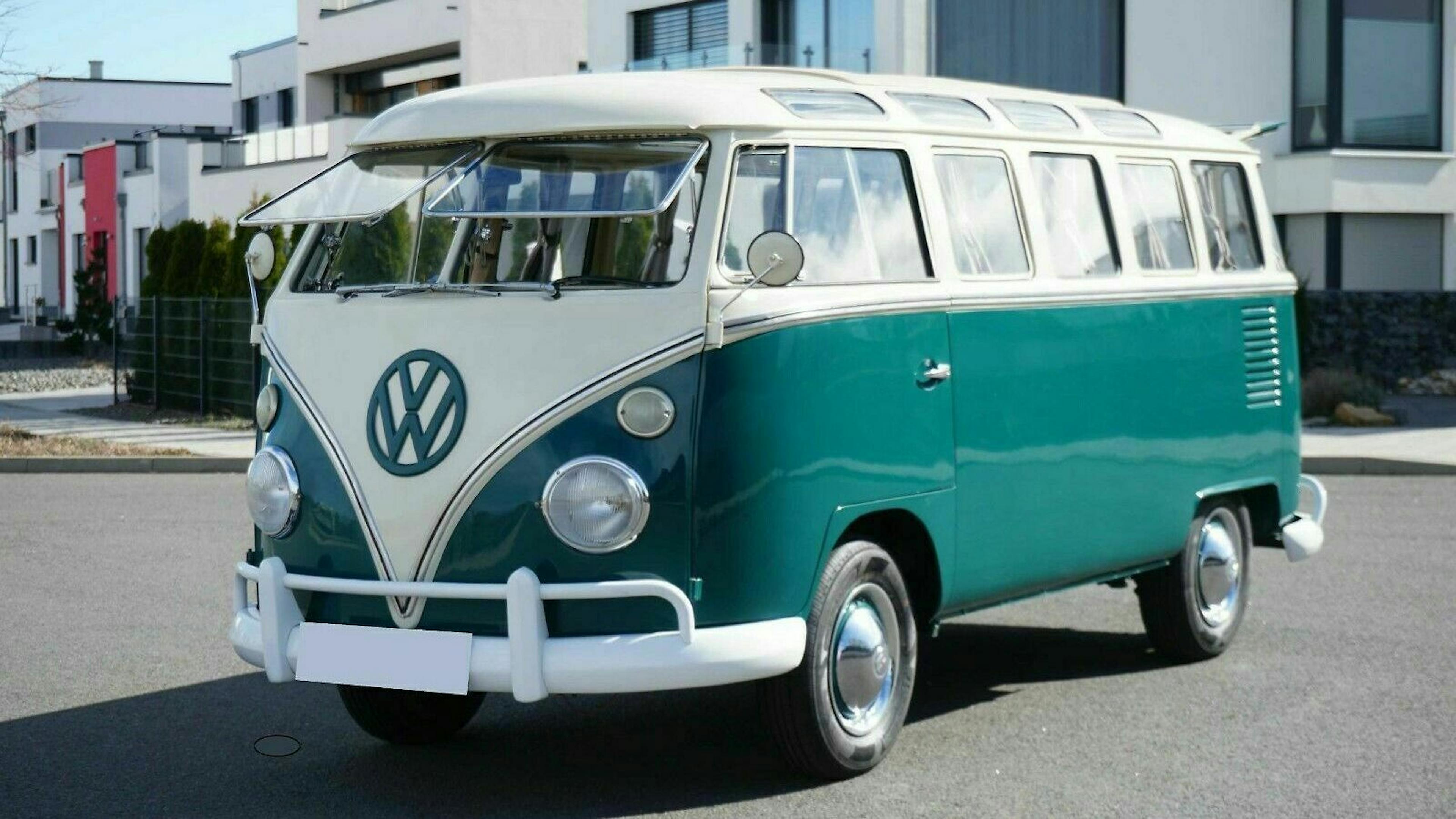 Ein türkis-weißer VW-Bus, Modell T1, steht auf einer Straße vor modernen Häusern. Klassisches Design mit runden Scheinwerfern und VW-Logo.