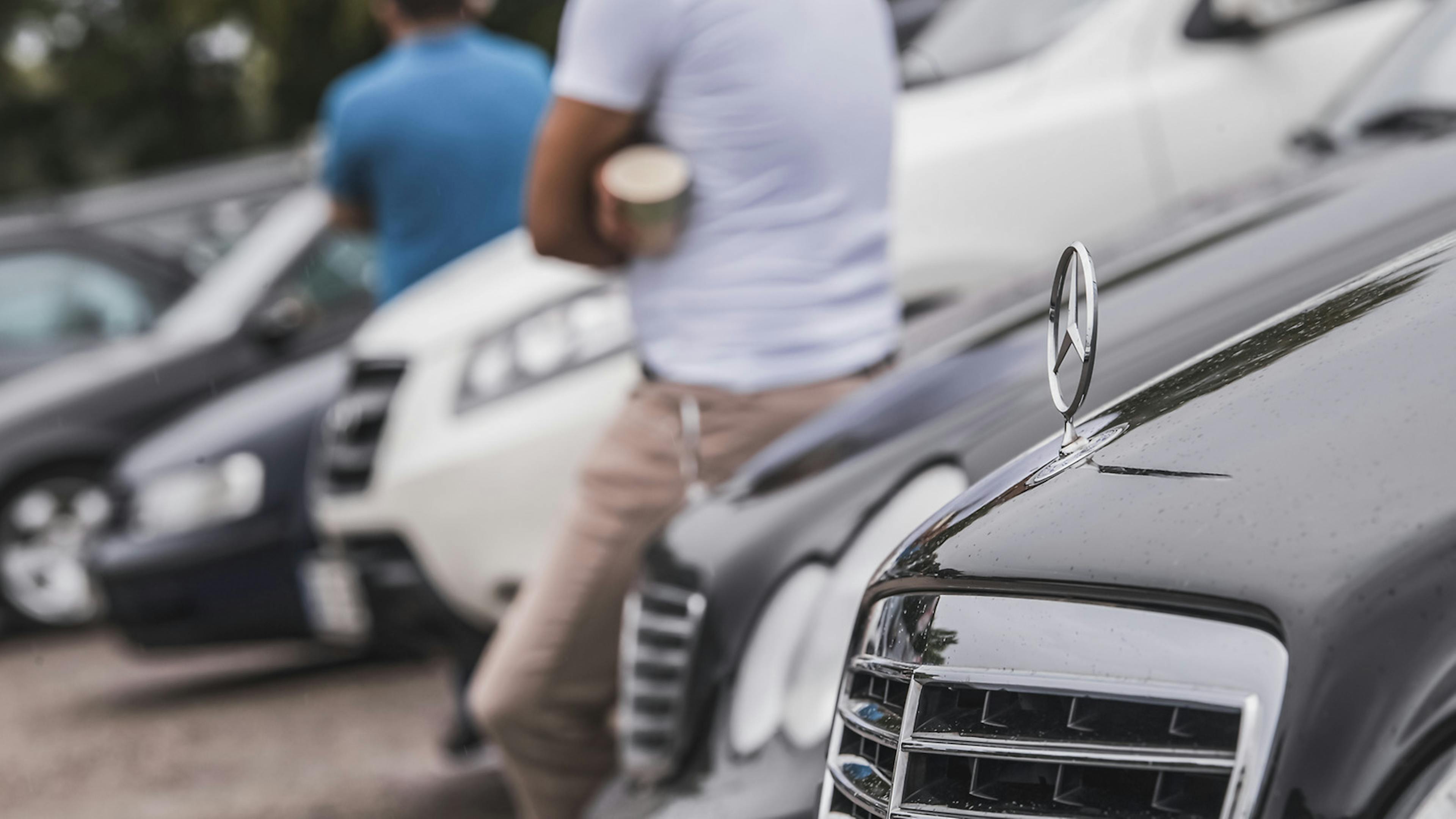 Autos stehen in einer Reihe, scharf im Vordergrund ragt ein Mercedes-Stern über dem Kühlergrill
