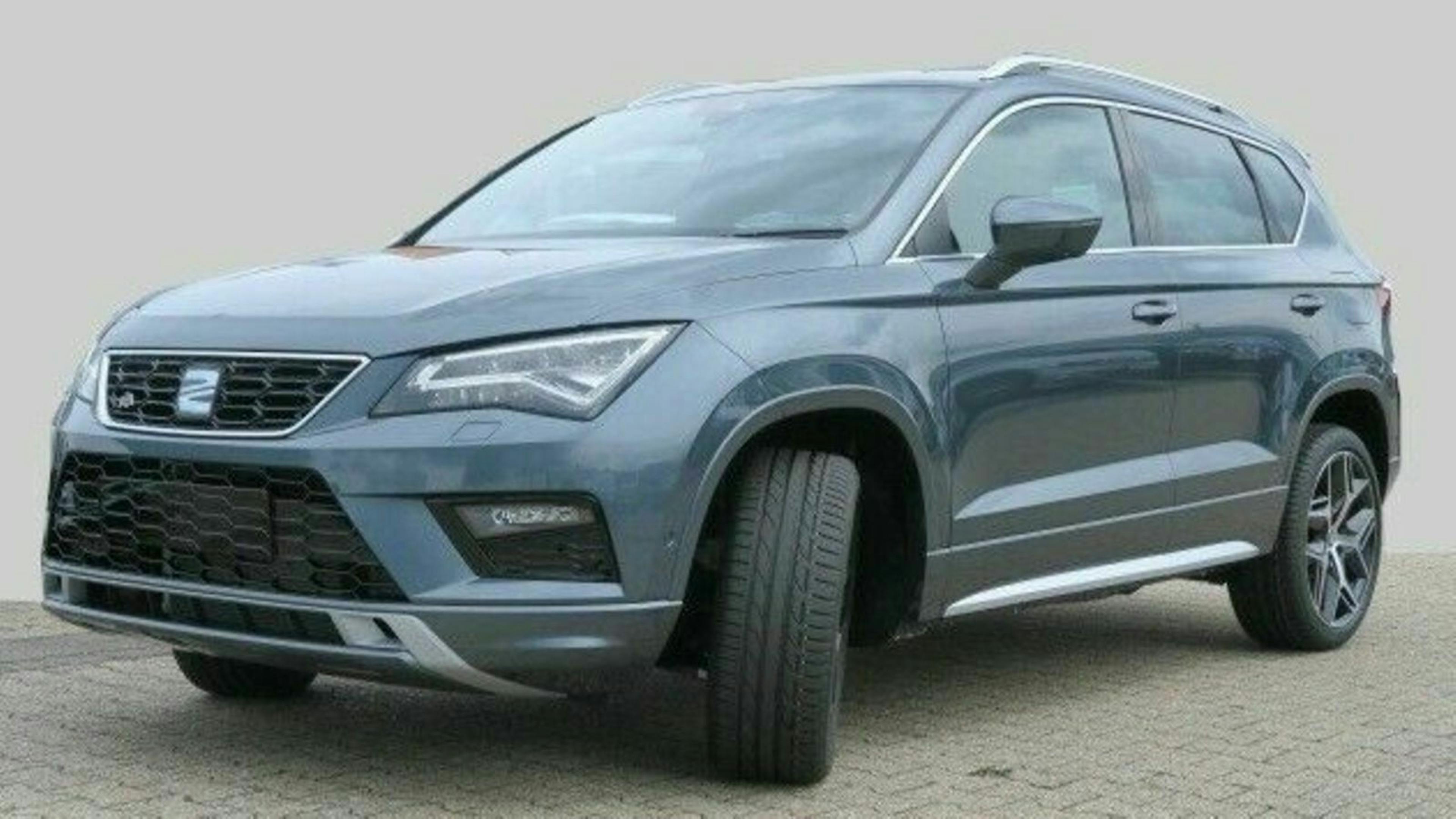 Grauer SUV der Marke SEAT Ateca, geparkt auf einem Pflastersteinboden vor grauem Hintergrund, Front- und Seitenansicht.