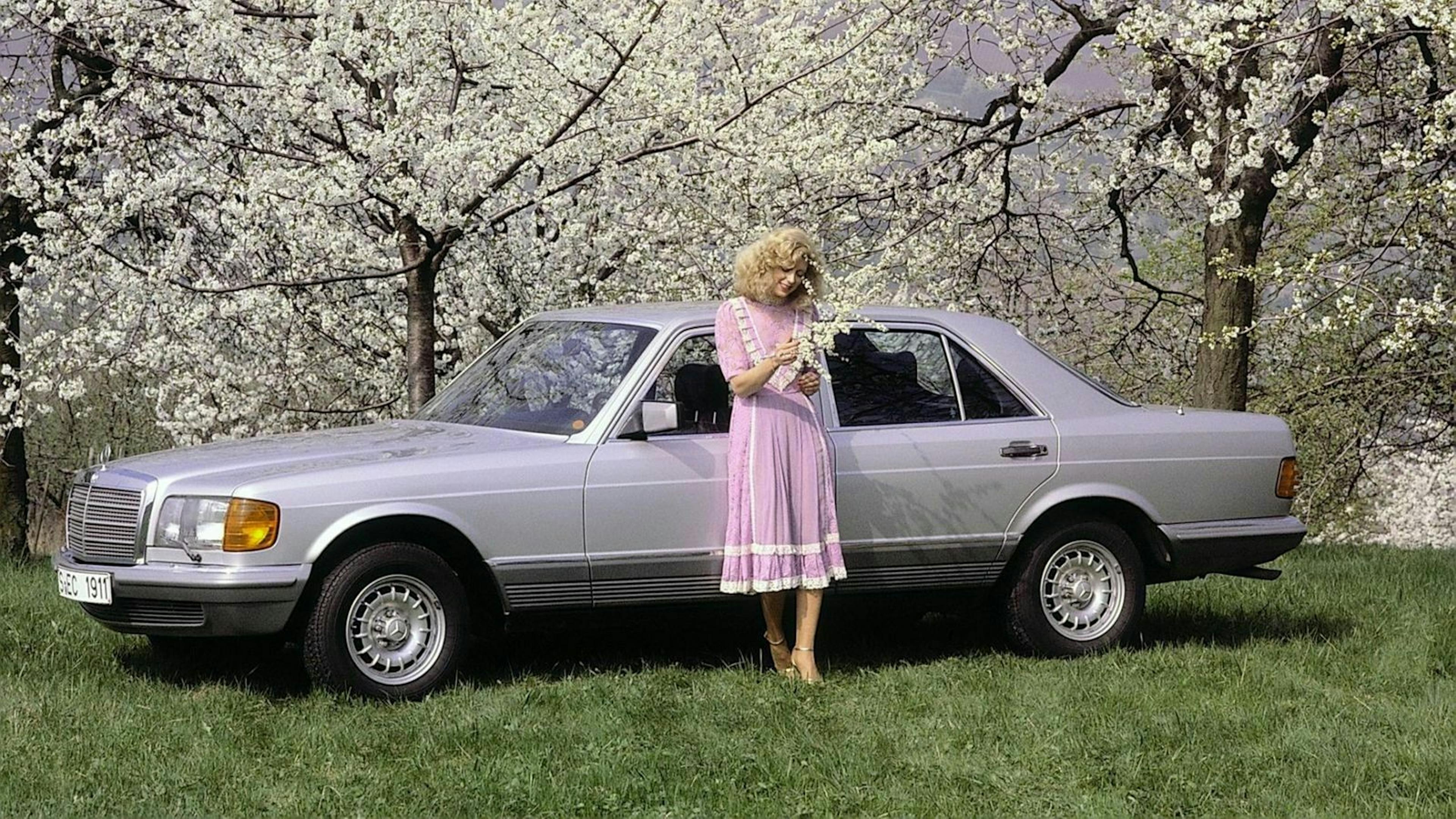 Frau in rosa Kleid steht vor einem silbernen Oldtimer-Auto, umgeben von blühenden Bäumen auf einer Wiese.