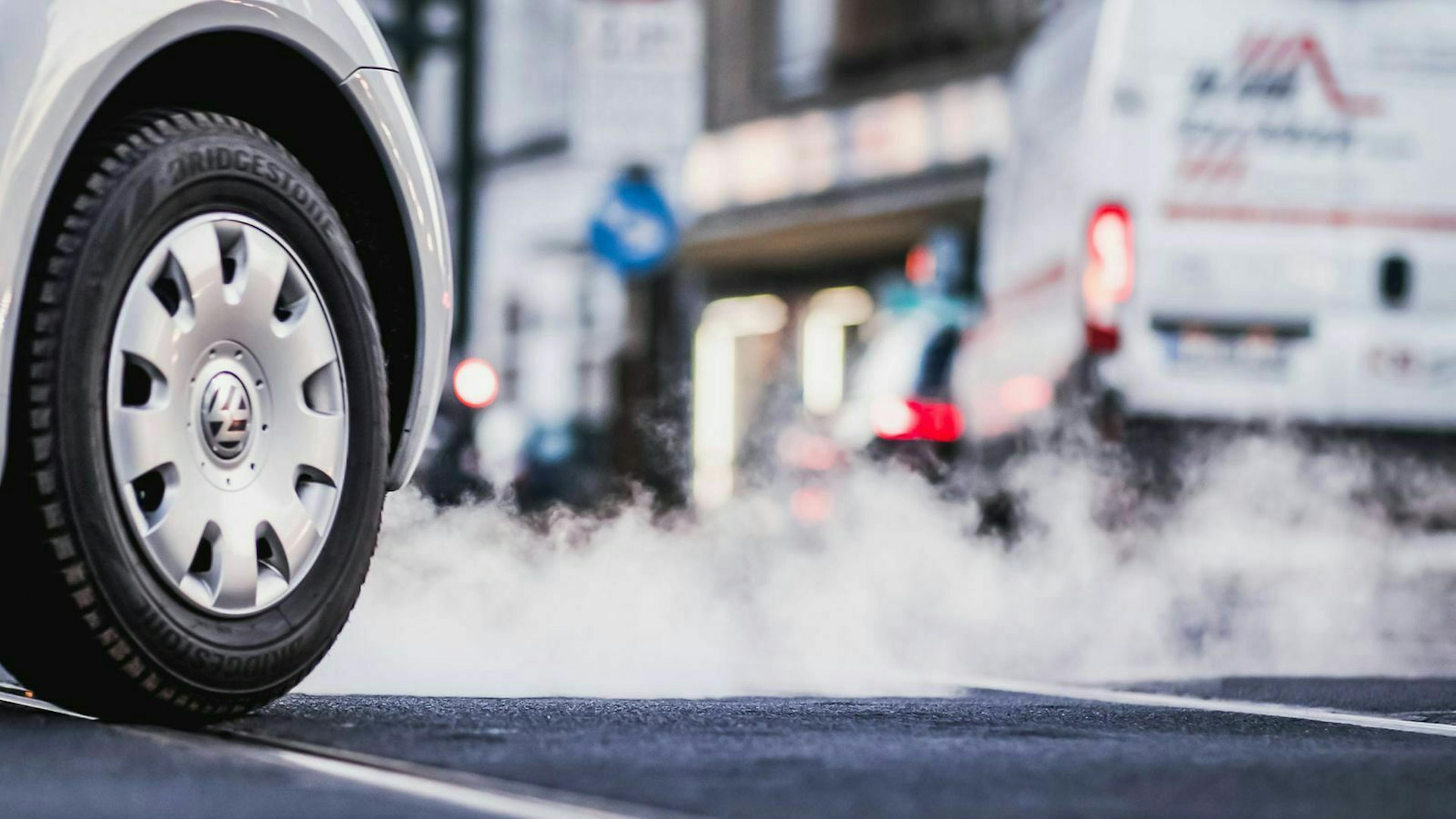 Im Vordergrund ist der Vorderreifen eines VW Golf zu sehen, im Hintergrund Abgase und unschärfer ein Transporter in einer Stadt.