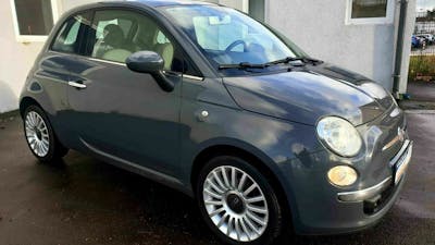 Ein schwarzer Fiat 500 mit Panoramadach steht vor einem weißen Gebäude.