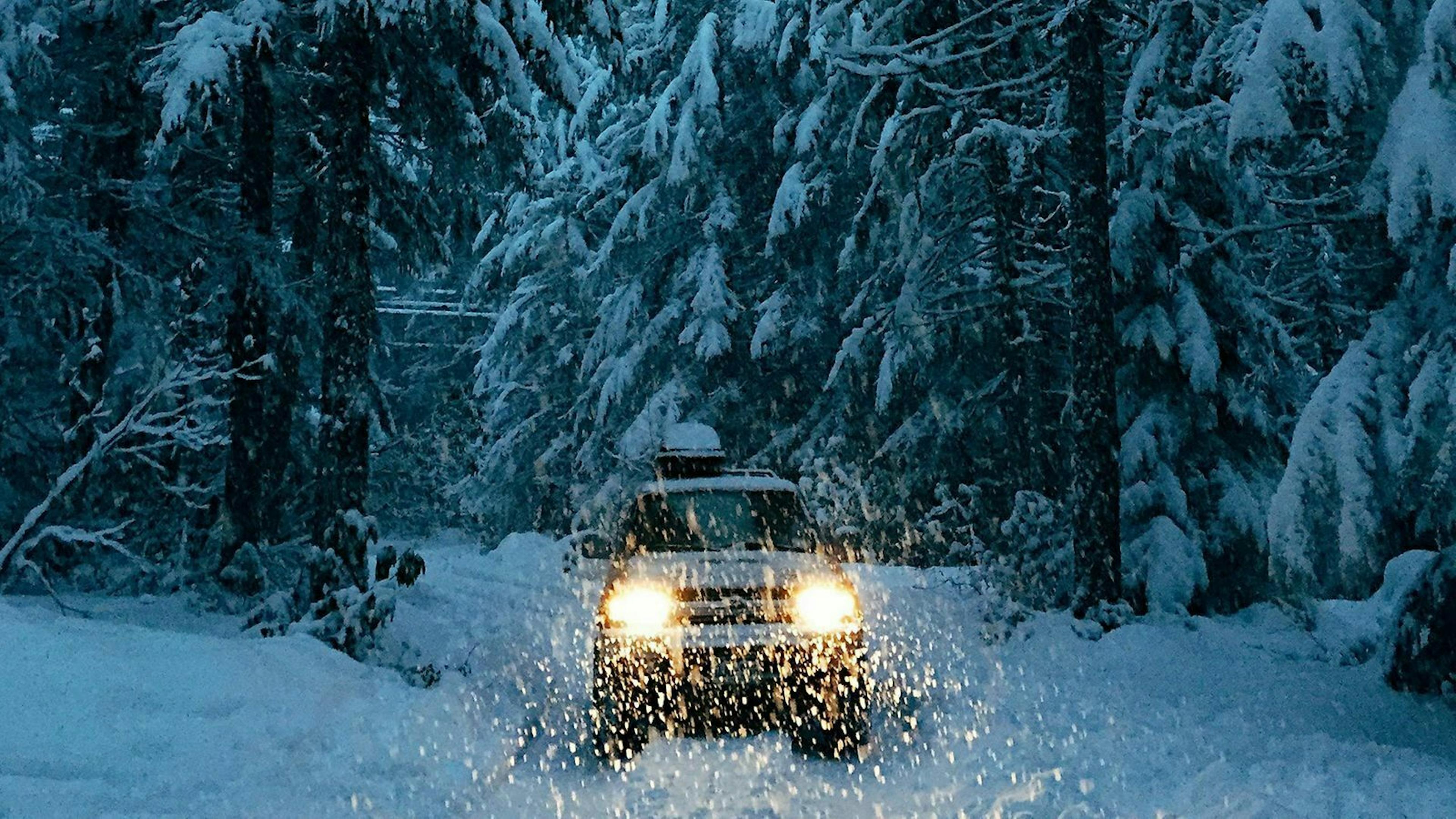 Ein Geländewagen fährt im Schneegestöber durch eine Winterlandschaft. Es dämmert und die Scheinwerfer leuchten den Weg.
