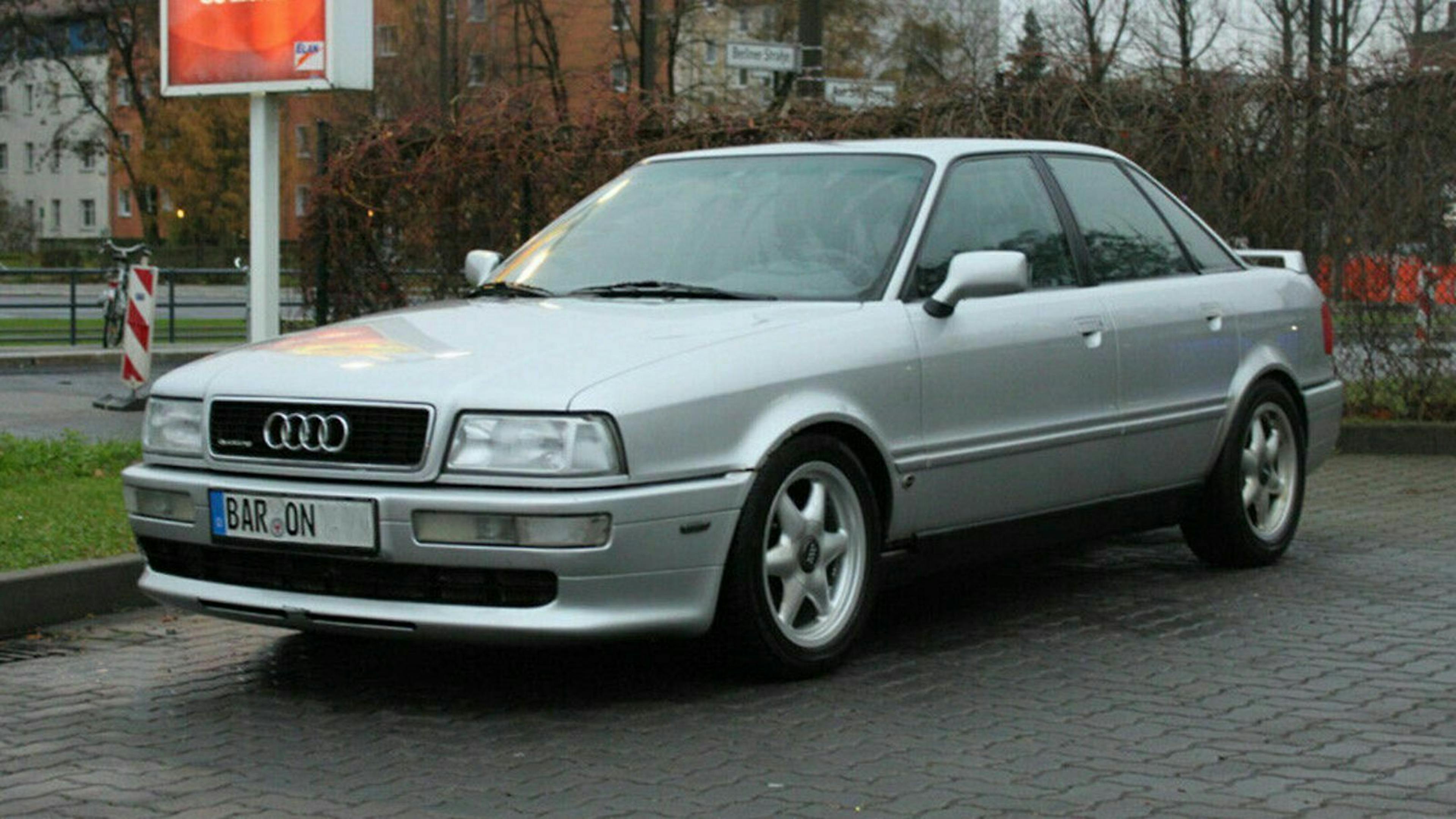 Silberner Audi mit deutschem Kennzeichen steht auf einem Parkplatz, Hintergrund mit Büschen und Gebäuden.