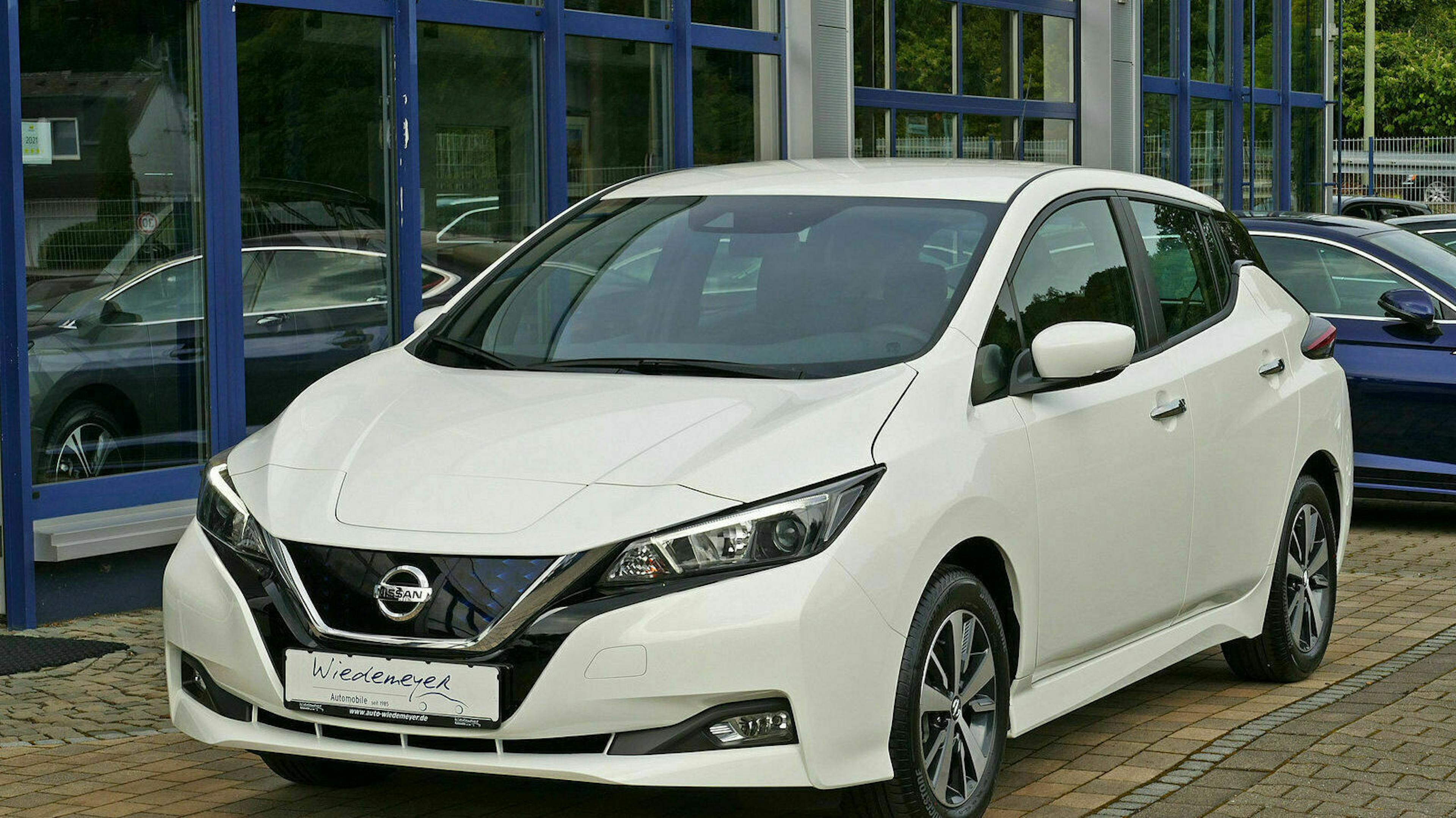 Ein weißer Nissan Leaf steht auf einem Parkplatz vor einem Gebäude mit bodentiefen Fenstern