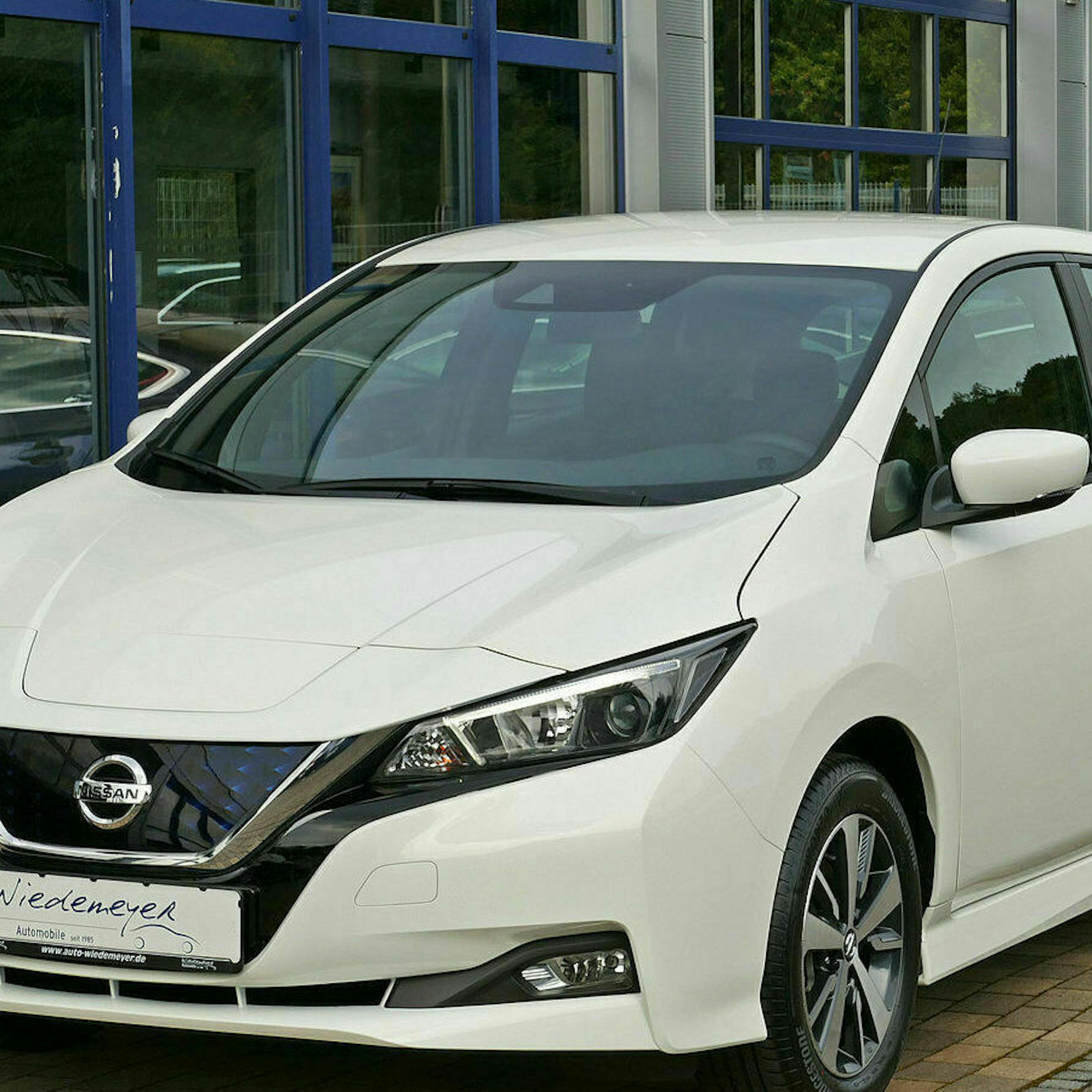 Ein weißer Nissan Leaf steht auf einem Parkplatz vor einem Gebäude mit bodentiefen Fenstern
