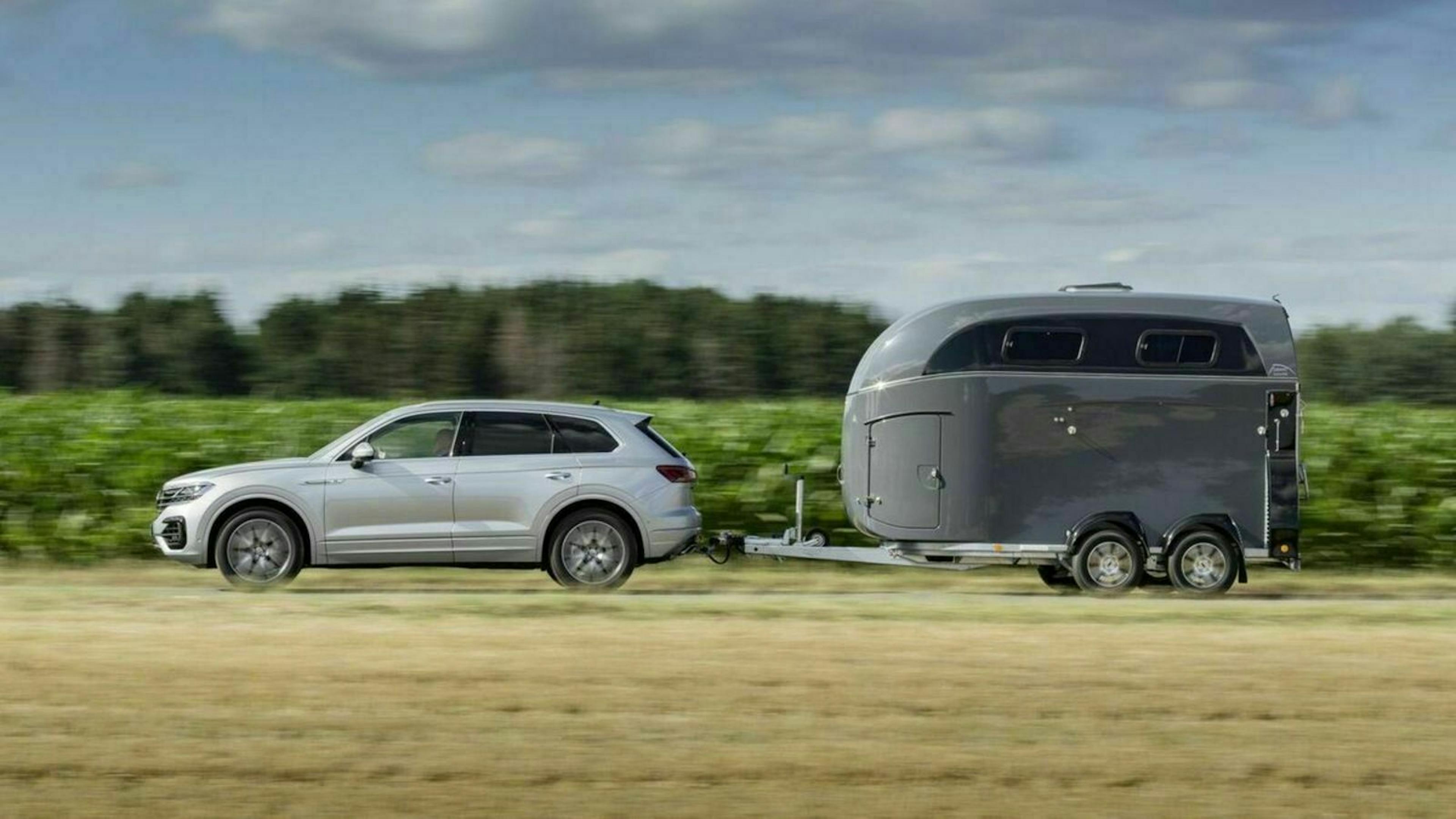 Ein silberner PKW mit einem grauen Pferdeanhänger fährt eine Landstraße entlang an einem Feld vorbei