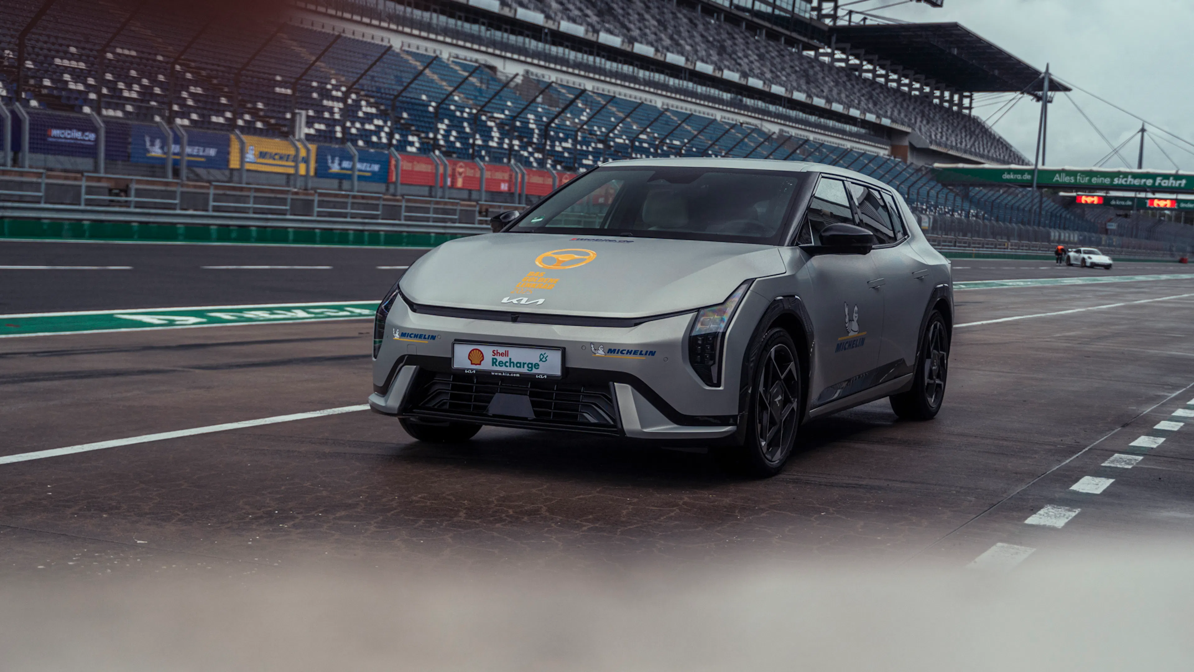 Grauer Kia EV4 in Dreiviertel-Frontansicht auf einer Rennstrecke. Auf dem Fahrzeug sind unter anderem Michelin- und Shell-Recharge-Logos zu sehen; im Hintergrund befinden sich Tribünen.