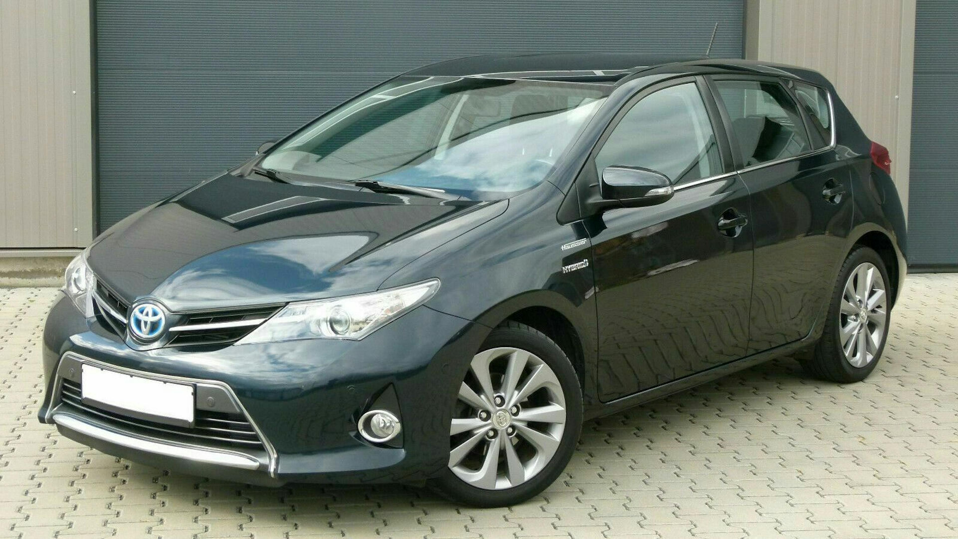 Dunkelgrauer Toyota Auris Hybrid, geparkt auf gepflastertem Boden vor einer grauen Wand. Fünftürige Karosserie mit modernen Scheinwerfern.