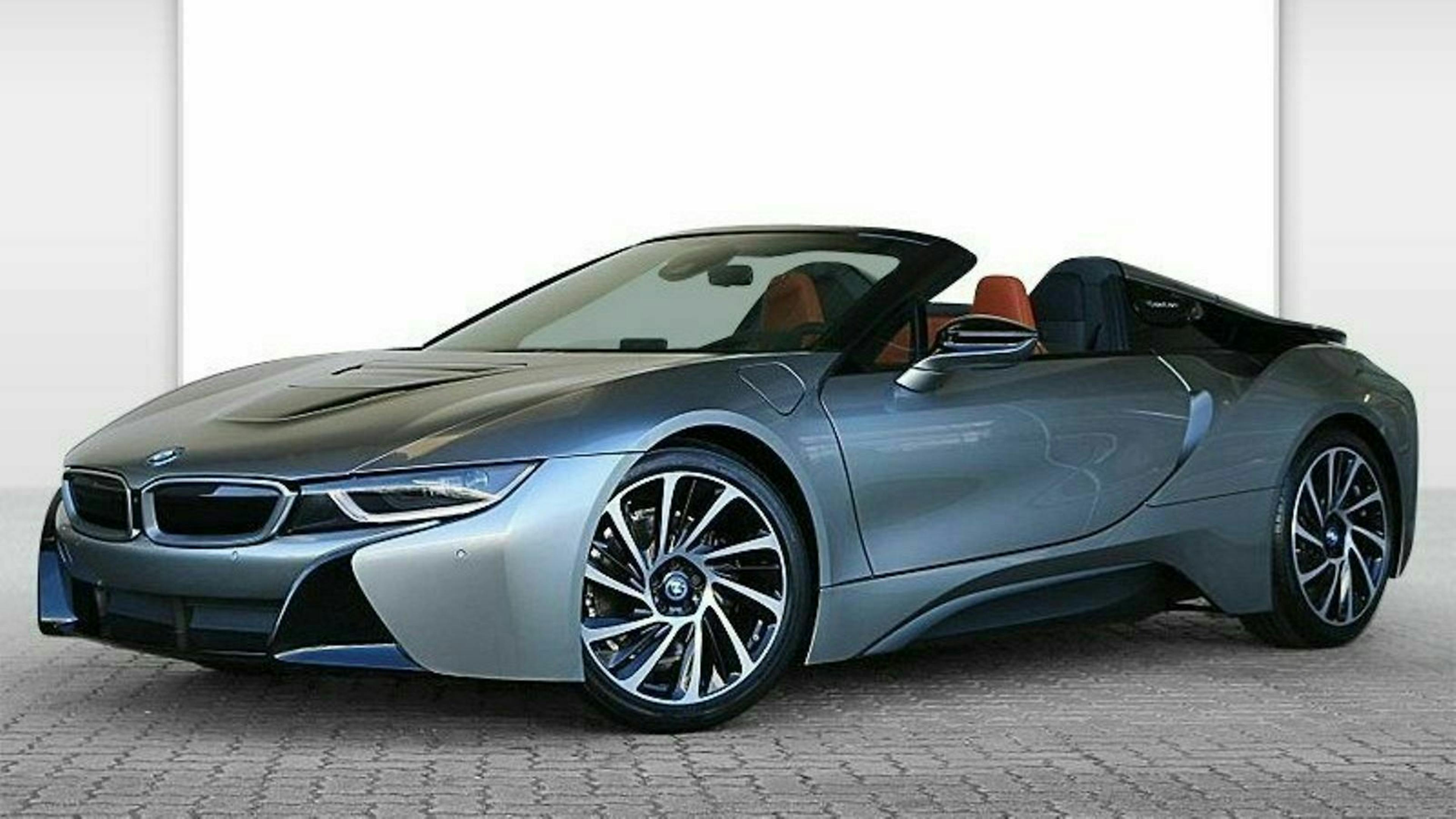 Grauer BMW i8 Roadster mit offenem Verdeck auf gepflastertem Boden. Futuristisches Design mit markanten Linien und sportlichem Erscheinungsbild.
