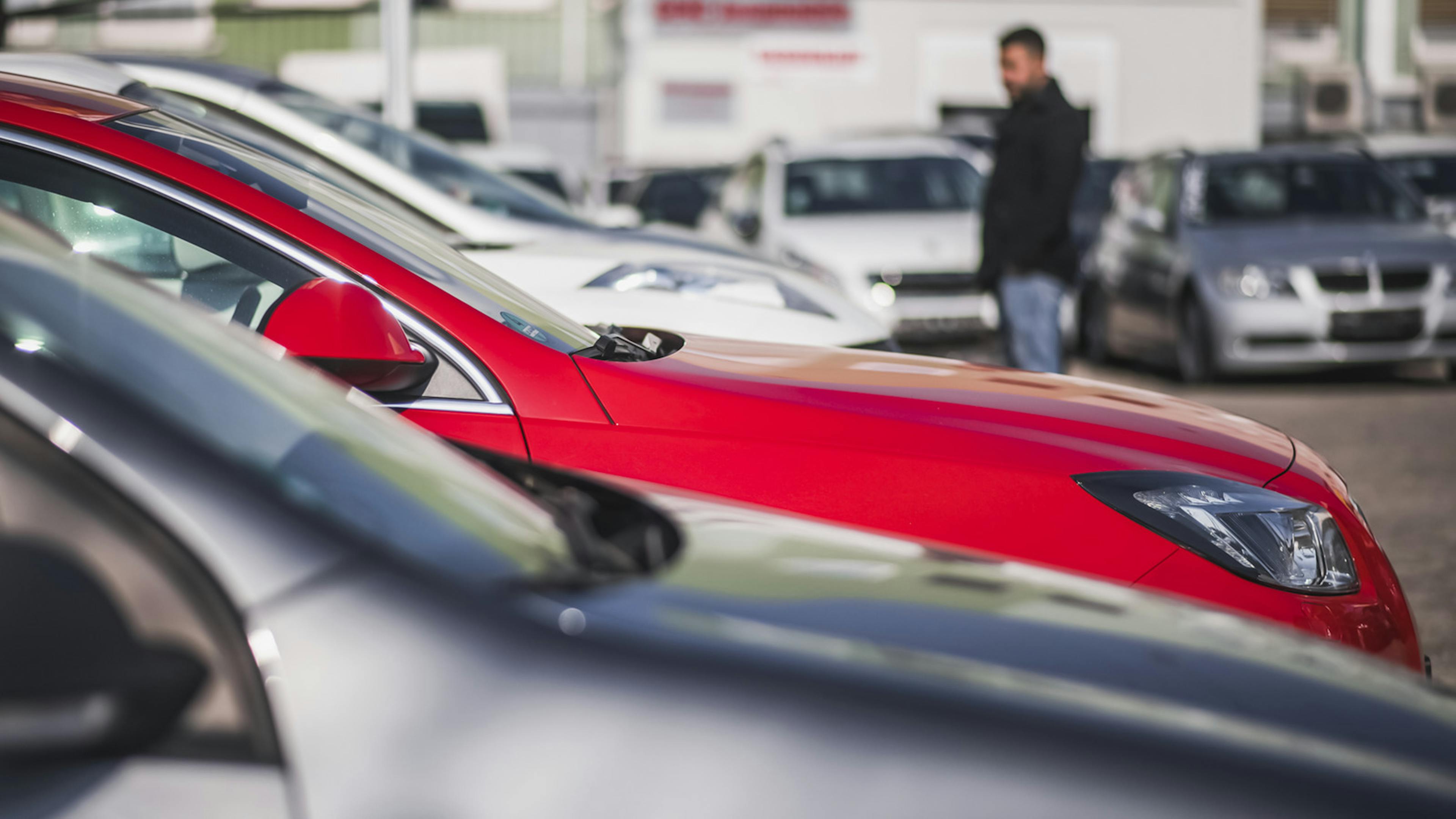 Ein roter Opel steht in einer Reihe mit anderen Autos, im Hintergrund unscharf schaut sich ein Mann Autos an.