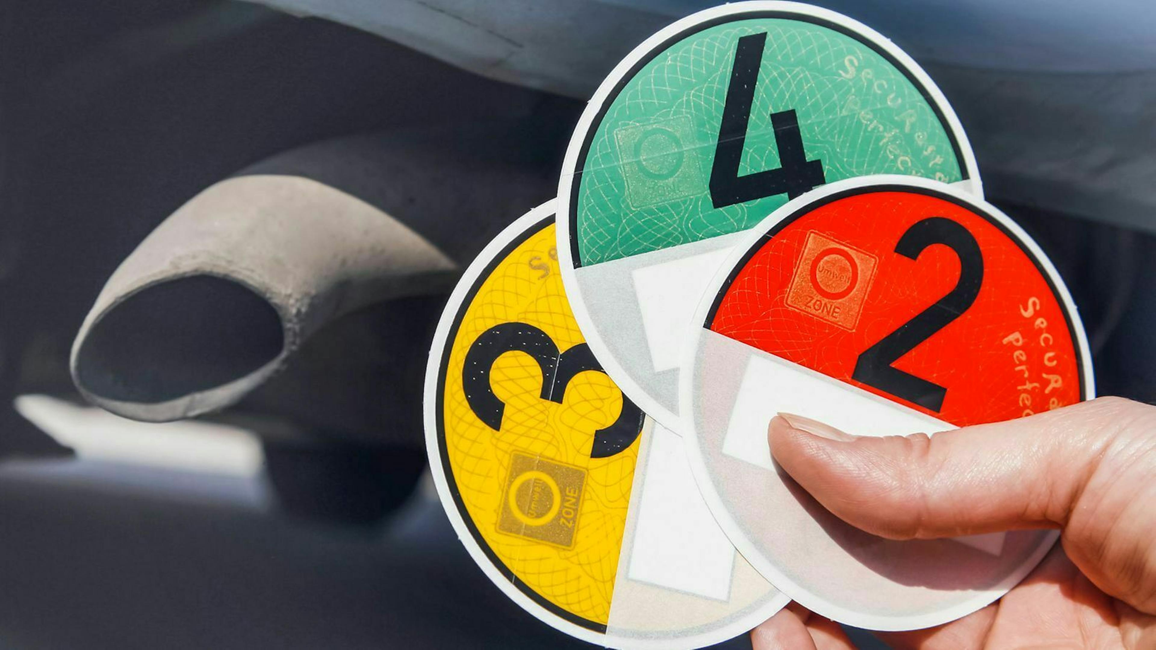 Jemand hält je eine gelbe, grüne und rote Umweltplakette vor einem PKW-Auspuff im Hintergrund.