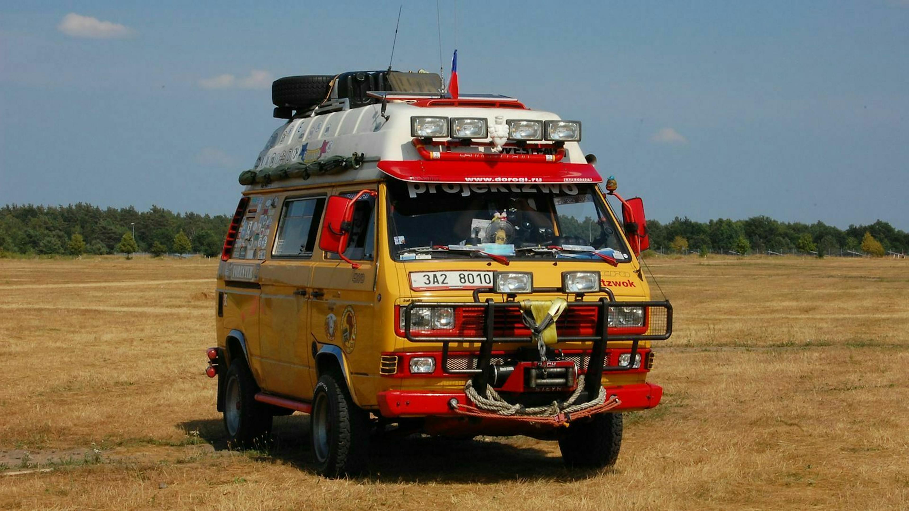 Ein VW Bulli mit Campingausstattung und Suchscheinwerfern fährt über eine Wiese.