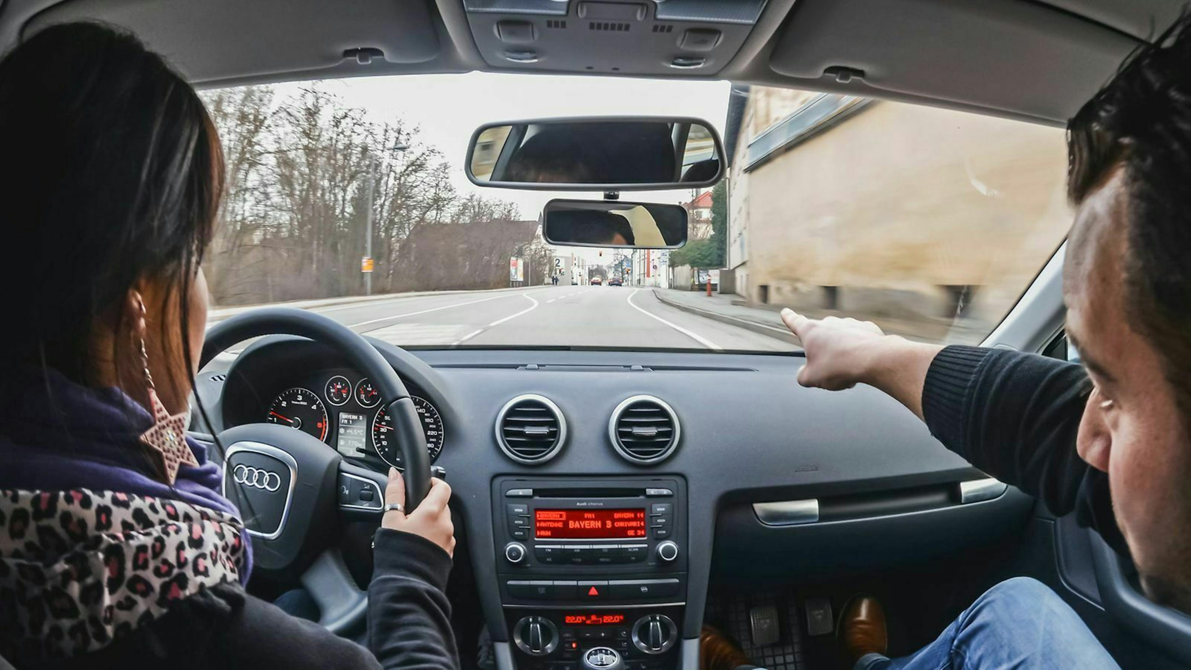 Eine junge Frau sitzt am Steuer eines Audi-A3-Fahrschulautos während sie vom Fahrlehrer auf dem Beifahrersitz instruiert wird.