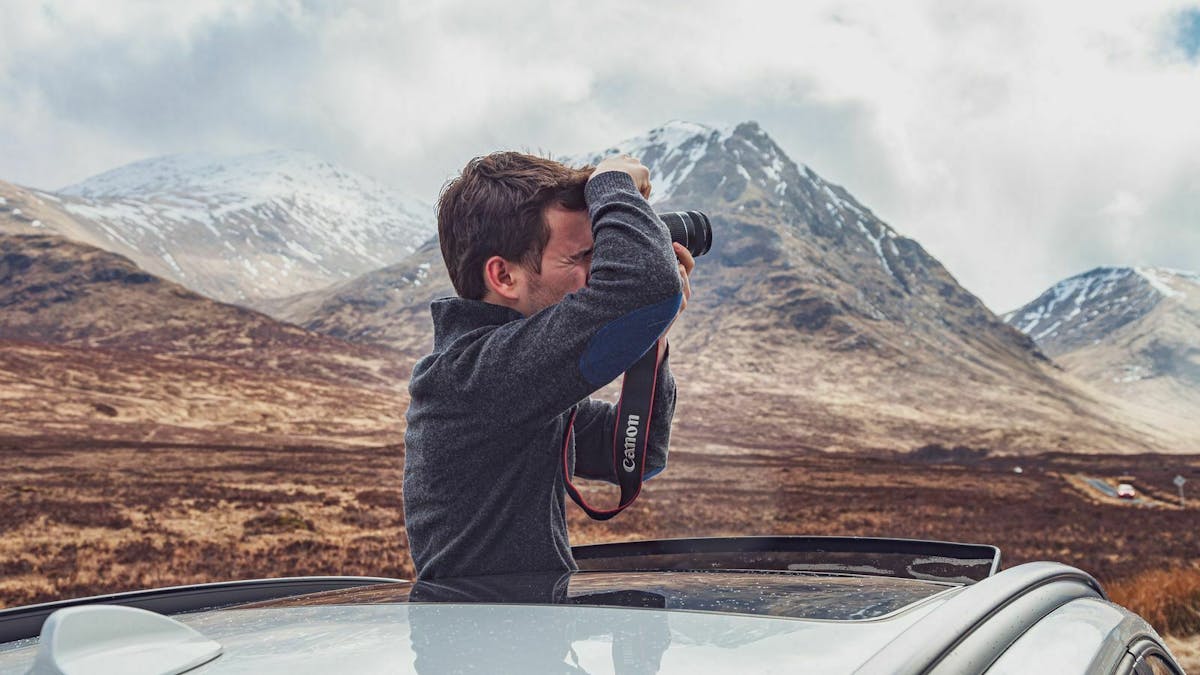Ein Mann schaut aus dem Schiebedach eines PKWs und fotografiert die Berglandschaft um ihn herum
