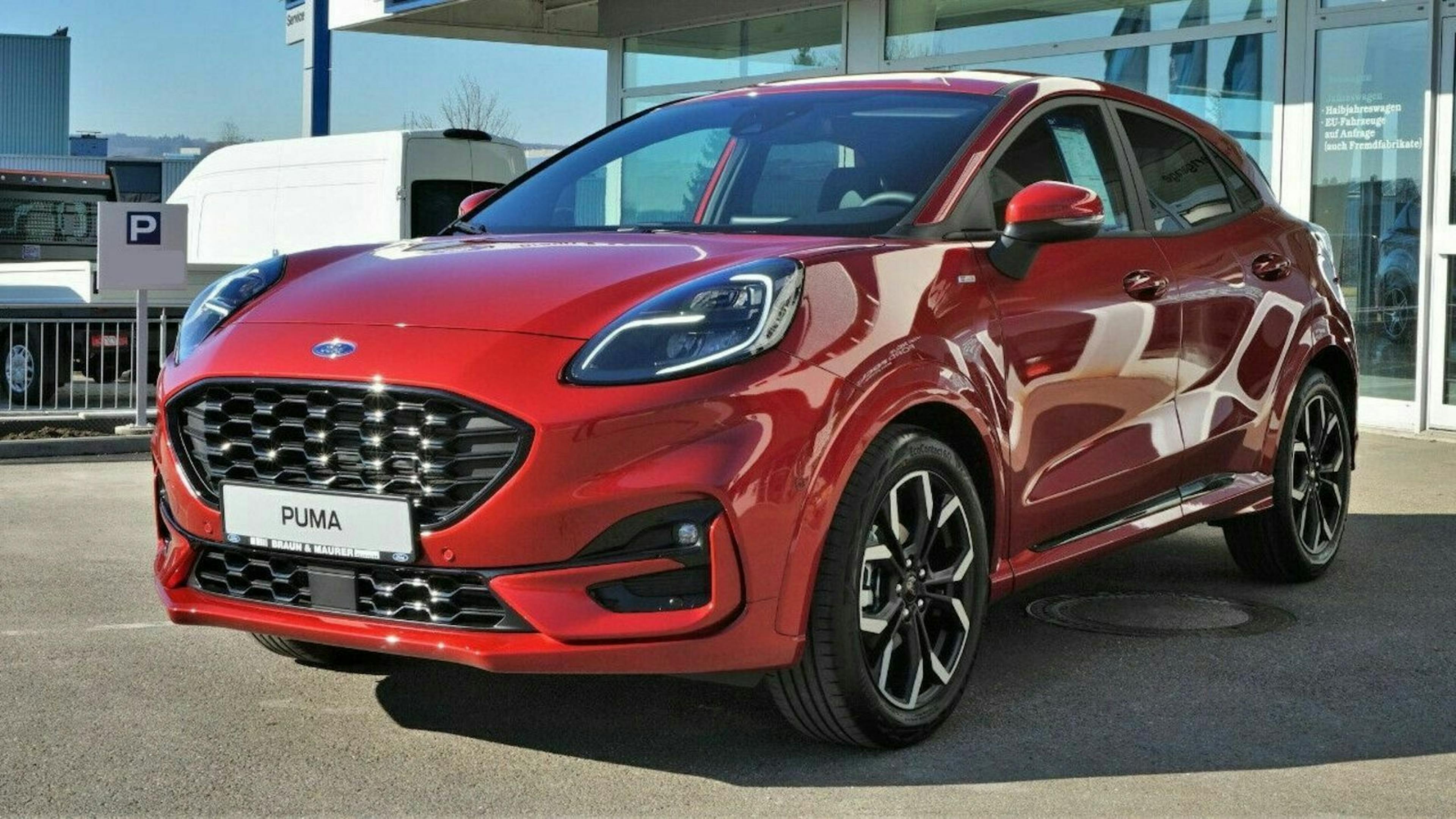 Roter Ford Puma in der Sonne, geparkt vor einem modernen Gebäude. Elegantes Design mit schwarzen Felgen und markantem Kühlergrill.