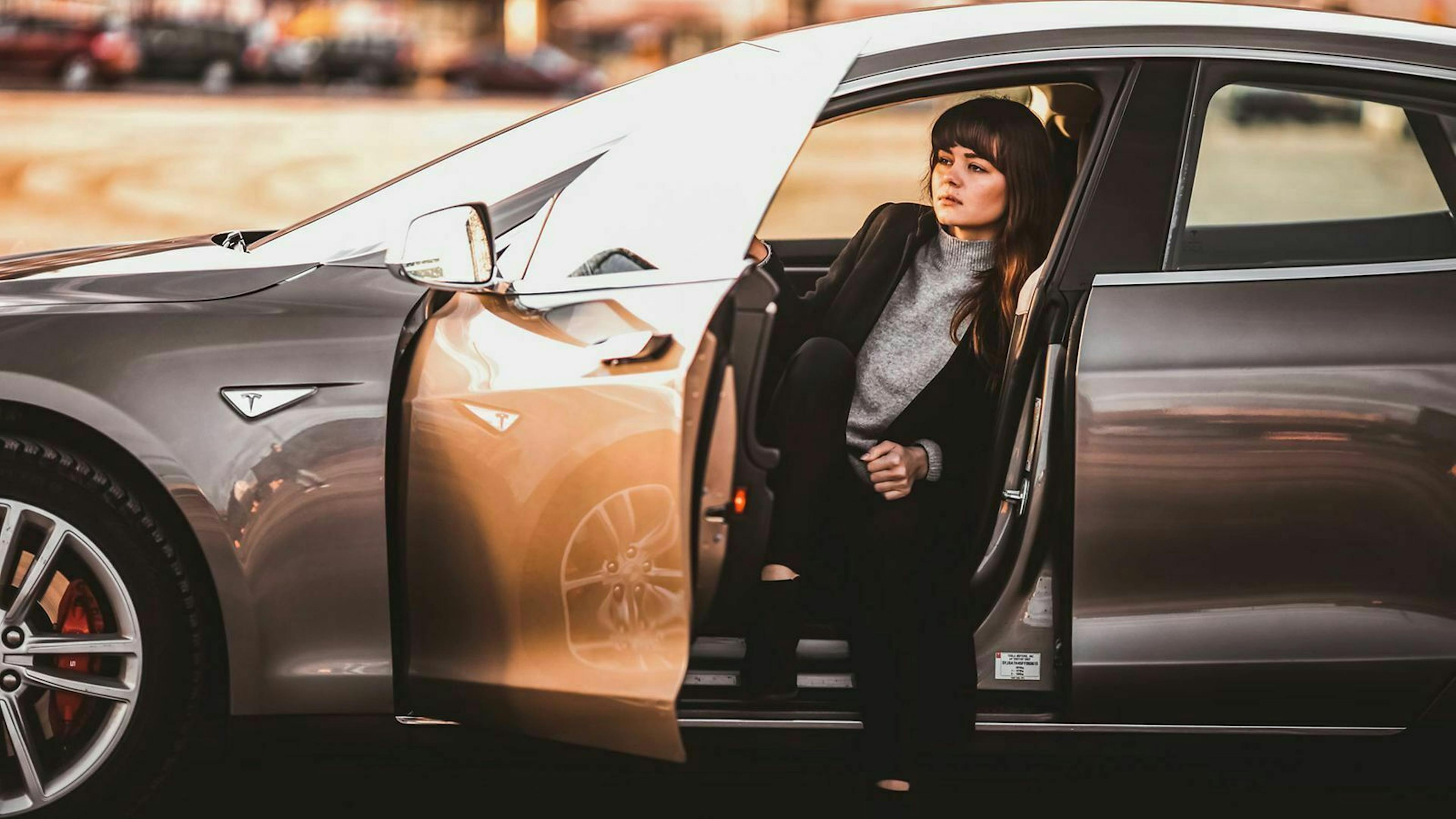 Eine junge Frau sitzt bei geöffneter Tür auf dem Fahrersitz einer grauen Limousine.