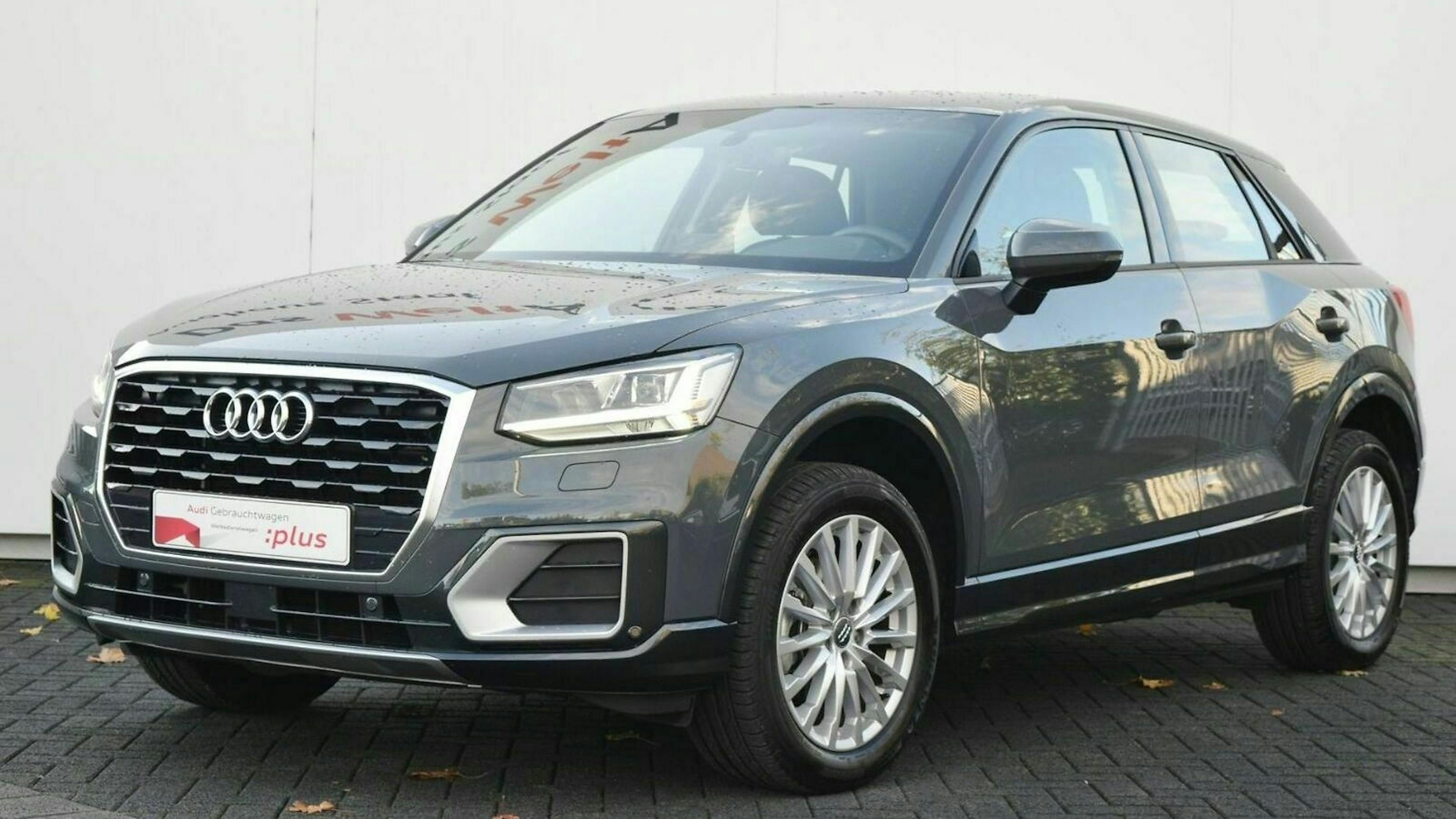 Grauer Audi Q2 SUV, seitlich von vorne zu sehen, auf einem gepflasterten Untergrund vor einer weißen Wand.