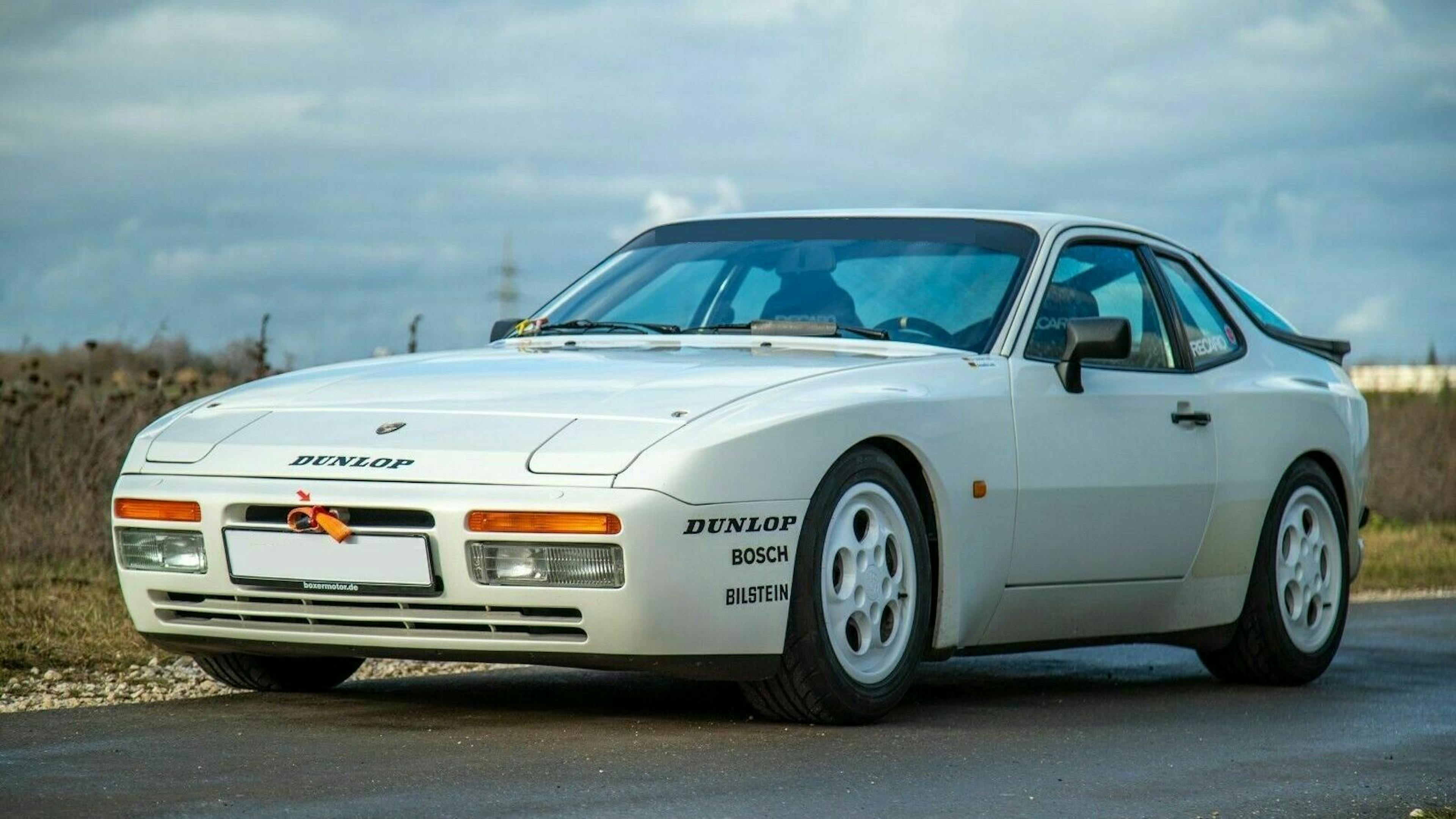 Weißer Sportwagen mit Dunlop- und Bosch-Logos an einem bewölkten Tag auf einer Straße geparkt.