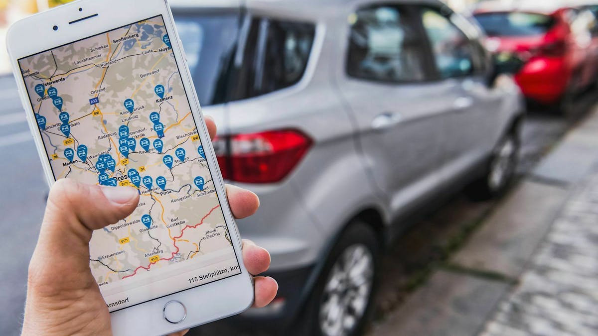 Jemand hält ein Smartphone, auf dem eine Park-App läuft. Unscharf im Hintergrund steht ein Auto.