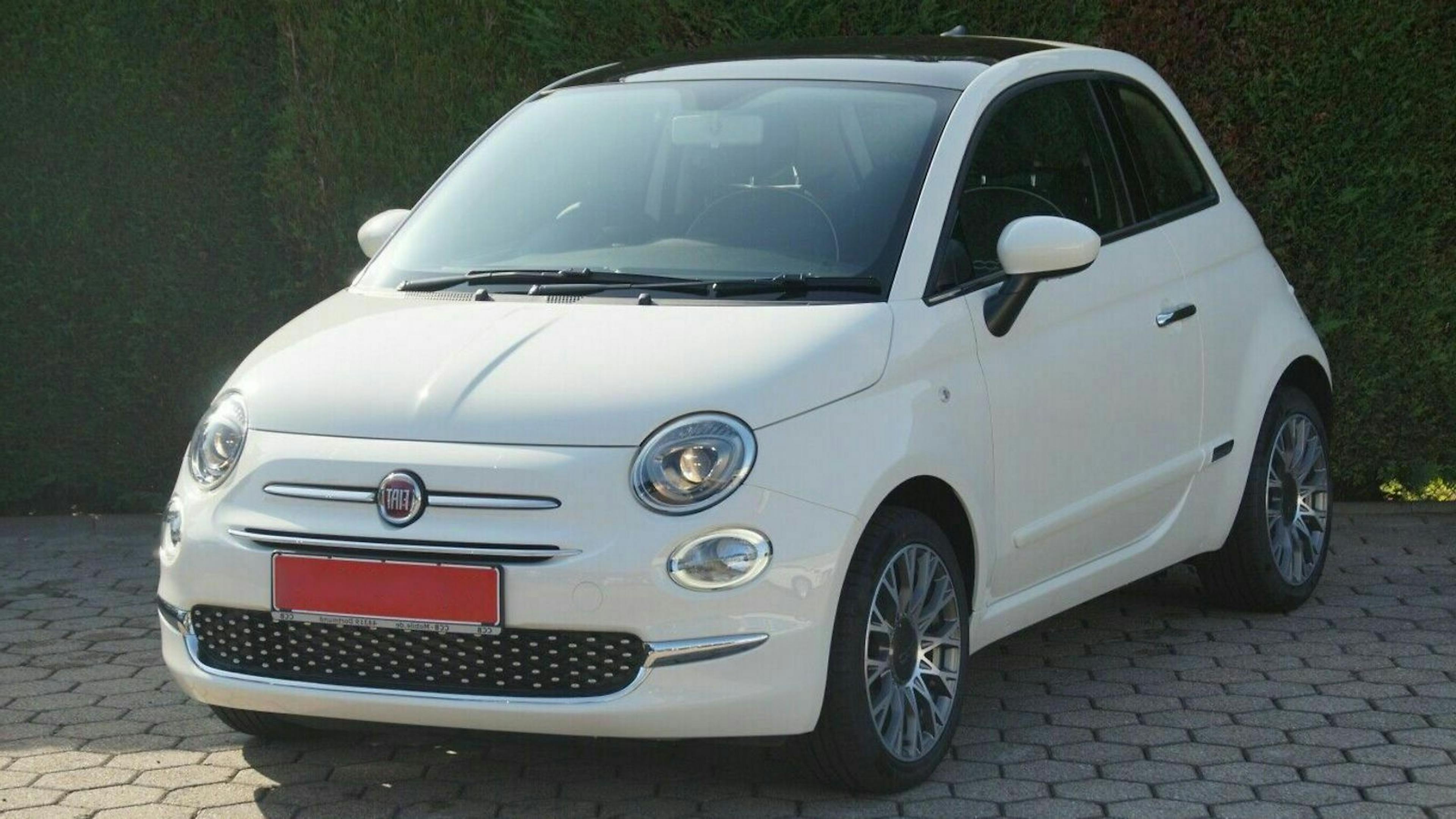 Weißer Fiat 500, steht auf gepflasterter Fläche. Kompaktwagen mit abgerundeten Formen. Hintergrund aus grünem Laubwerk.